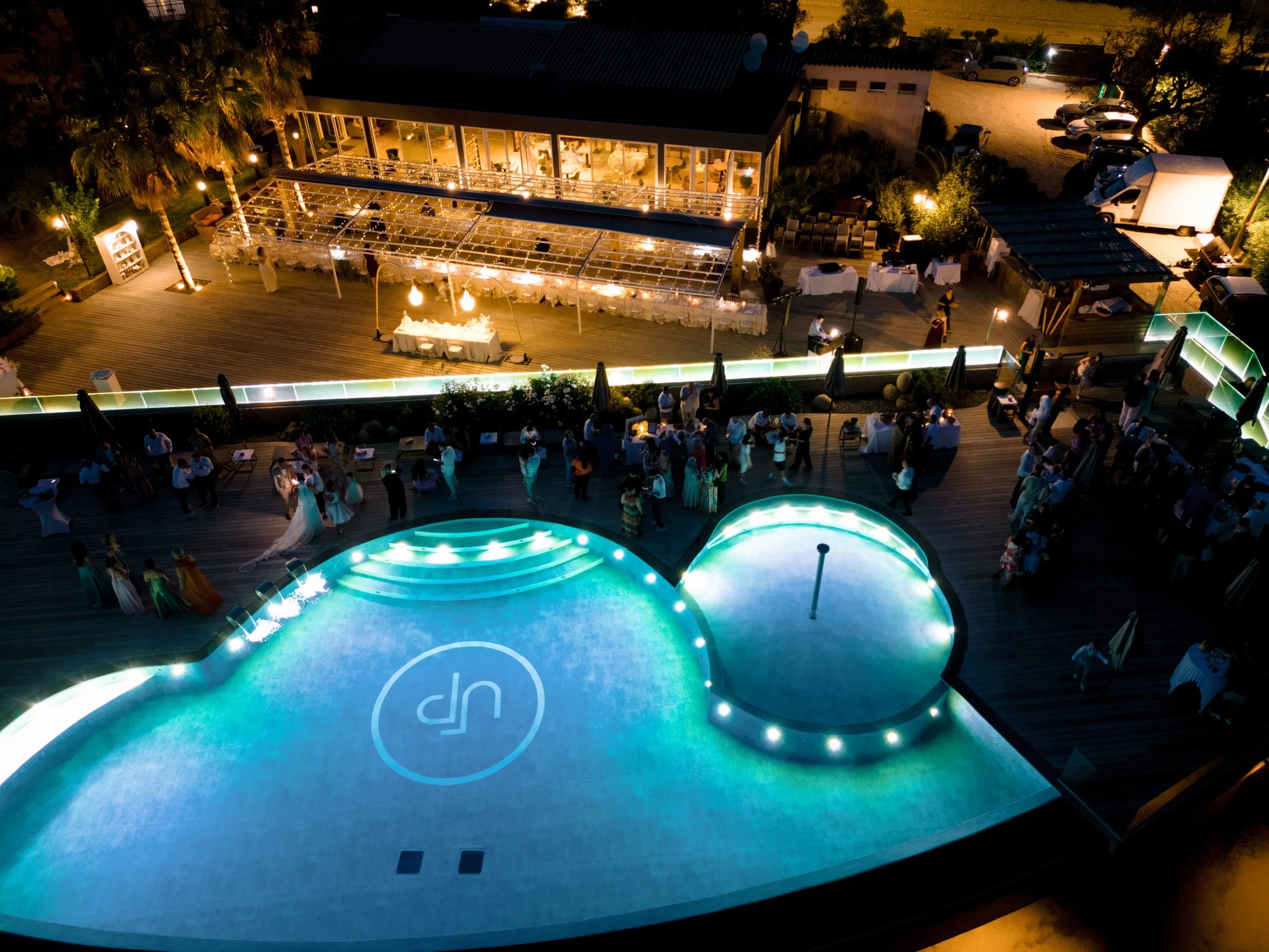 Vue aérienne d'une fête en soirée avec une piscine illuminée, un espace extérieur avec des personnes dansant, des tables avec des nappes blanches et des invités. Il y a un bâtiment avec une terrasse éclairée et un espace pour le banquet. L'ambiance e