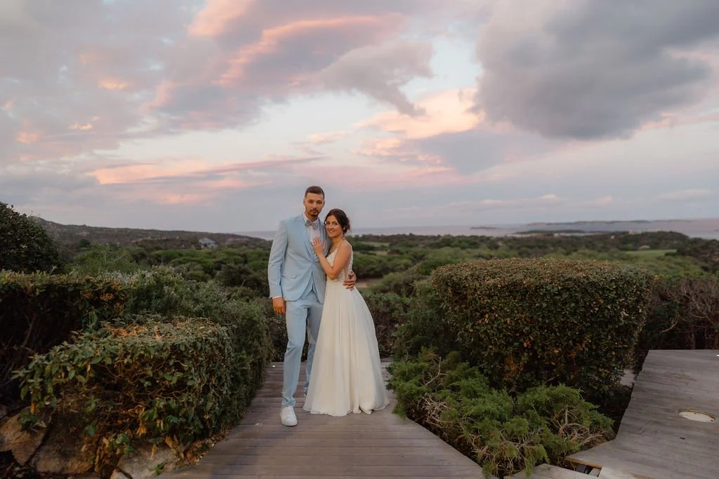 Un couple en robes de mariage se tenant par la taille sur un pont en bois, avec un paysage naturel au coucher du soleil en arrière-plan.