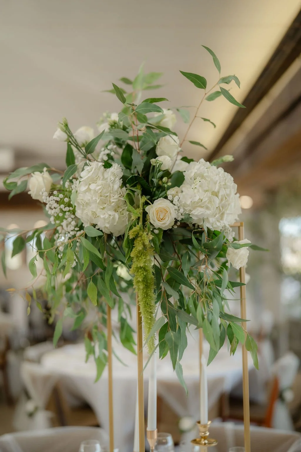 Centre de table floral avec boules de hortensias blanches, roses blanches, lisianthus et feuillage vert, avec des chandeliers dorés à bougies dans un cadre intérieur décoré pour un évènement