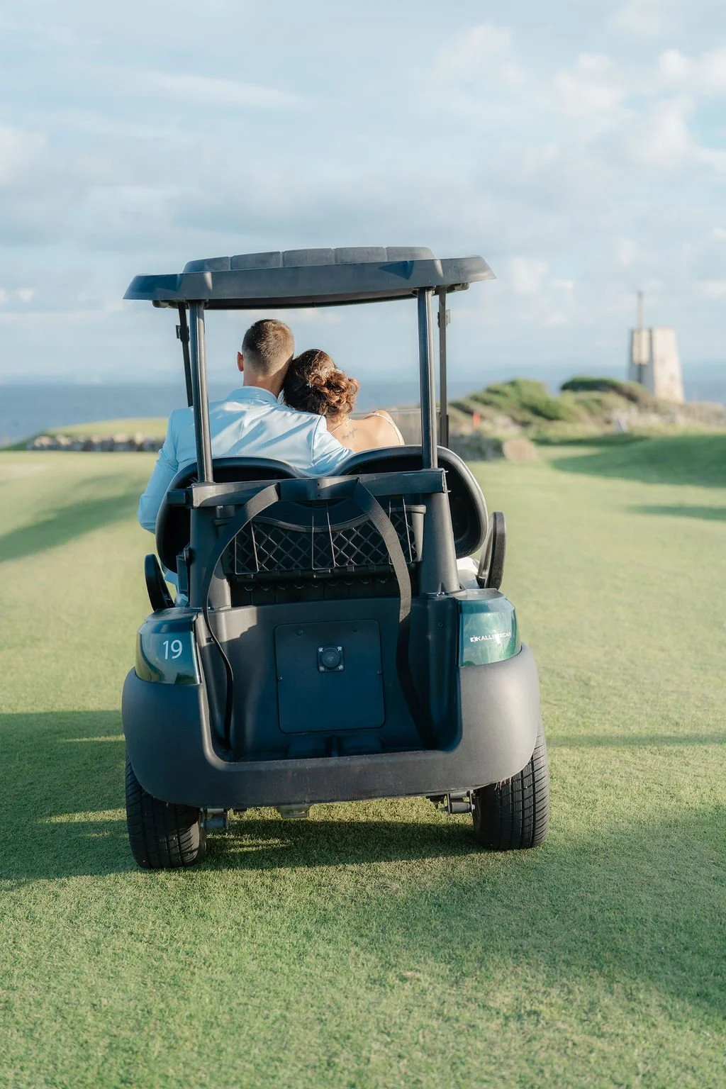 Un couple assis dans une voiturette de golf sur un parcours verdoyant, avec un château en pierre en arrière-plan sous un ciel partiellement nuageux.