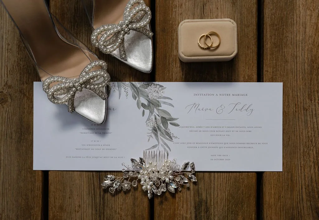 Des chaussures à talons avec des nœuds décoratifs en perles, un secret avec des alliances en or, une invitation de mariage, un peigne à cheveux décoratif avec des pierres précieuses, le tout placé sur une surface en bois.
