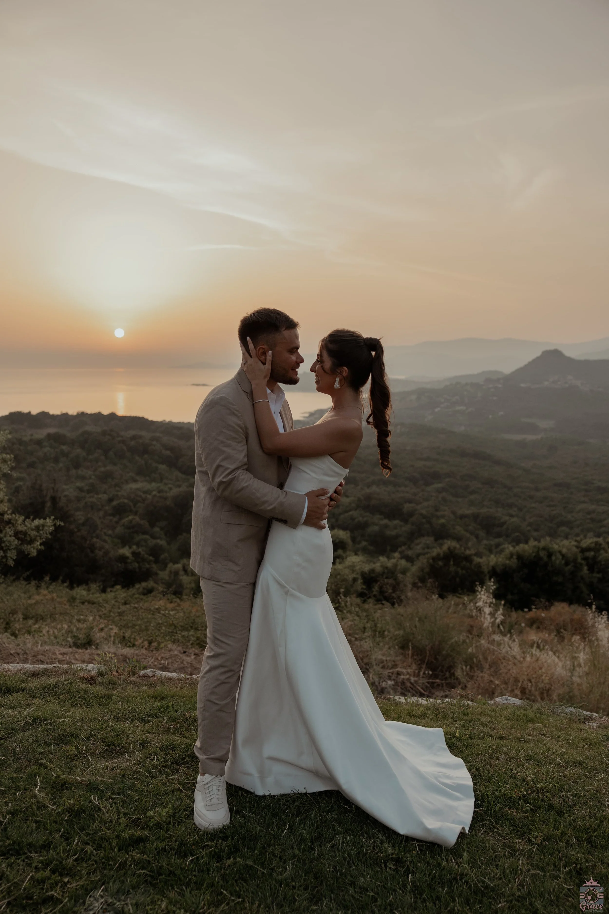 Un couple de mariés se tient lors d'un coucher de soleil dans un paysage verdoyant, avec des montagnes en arrière-plan et la mer au loin.
