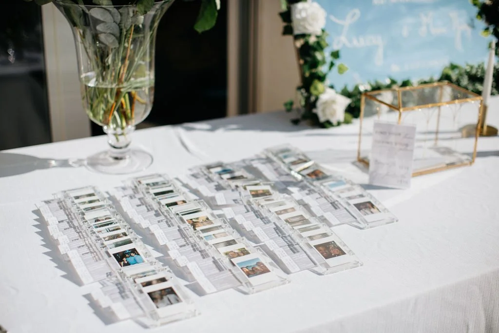 Une table blanche avec des bandes de photos Polaroid disposées en rangs, une grande vase avec des fleurs, un cadre en verre doré, et un tableau blanc avec des fleurs blanches en arrière-plan.