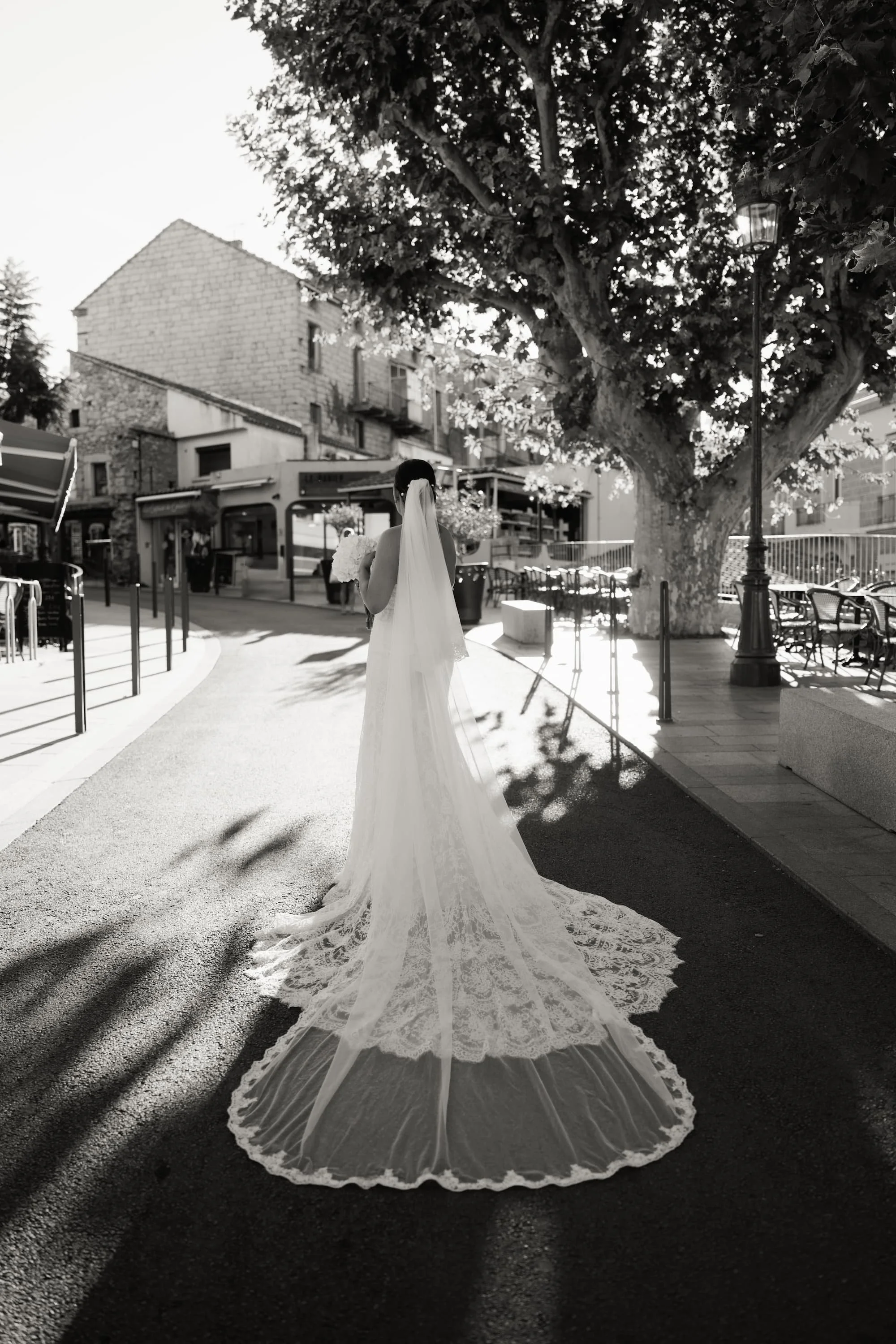 Une mariée en robe de mariée longue et voilée, vue de dos, se tenant devant un grand arbre, dans un espace urbain en plein air.