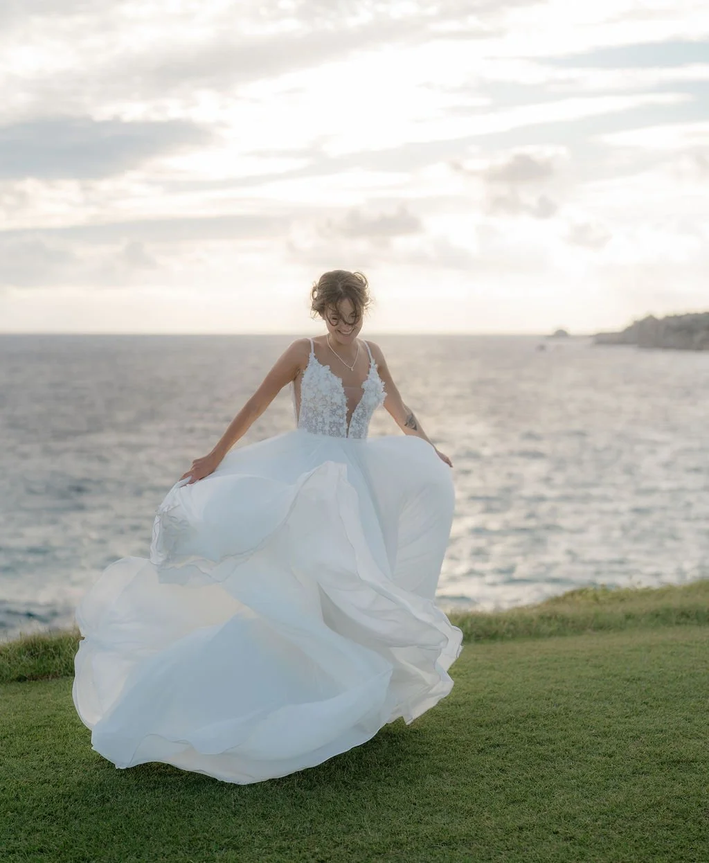 Jeune femme en robe de mariée blanches, dansant sur une pelouse près de la mer au coucher du soleil.