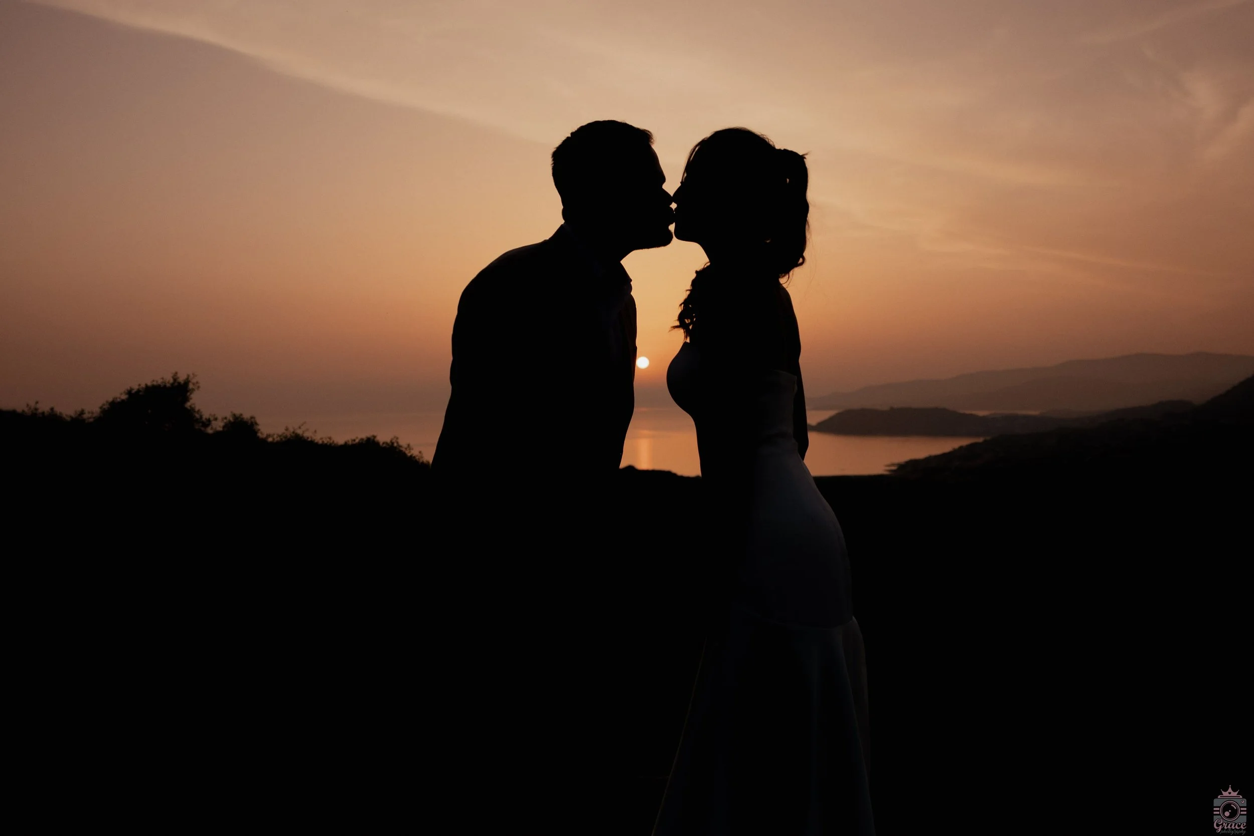Silhouette d'un couple s'embrassant au coucher du soleil, sur une scène extérieure avec un paysage de montagnes et d'eau en arrière-plan.