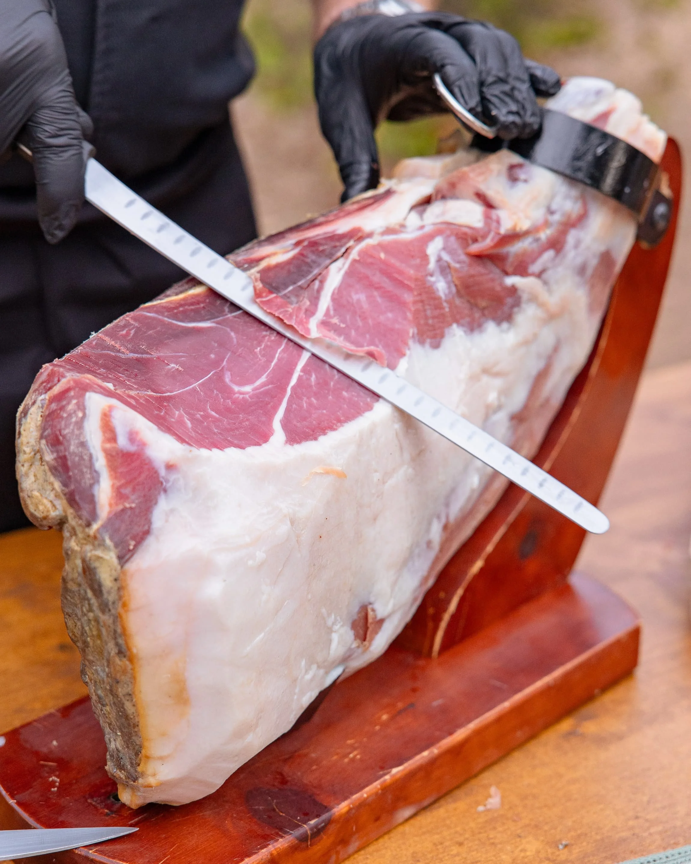 Une personne portant des gants noirs coupe un gros morceau de jambon avec un couteau à lame longue, le jambon repose sur une planche en bois.