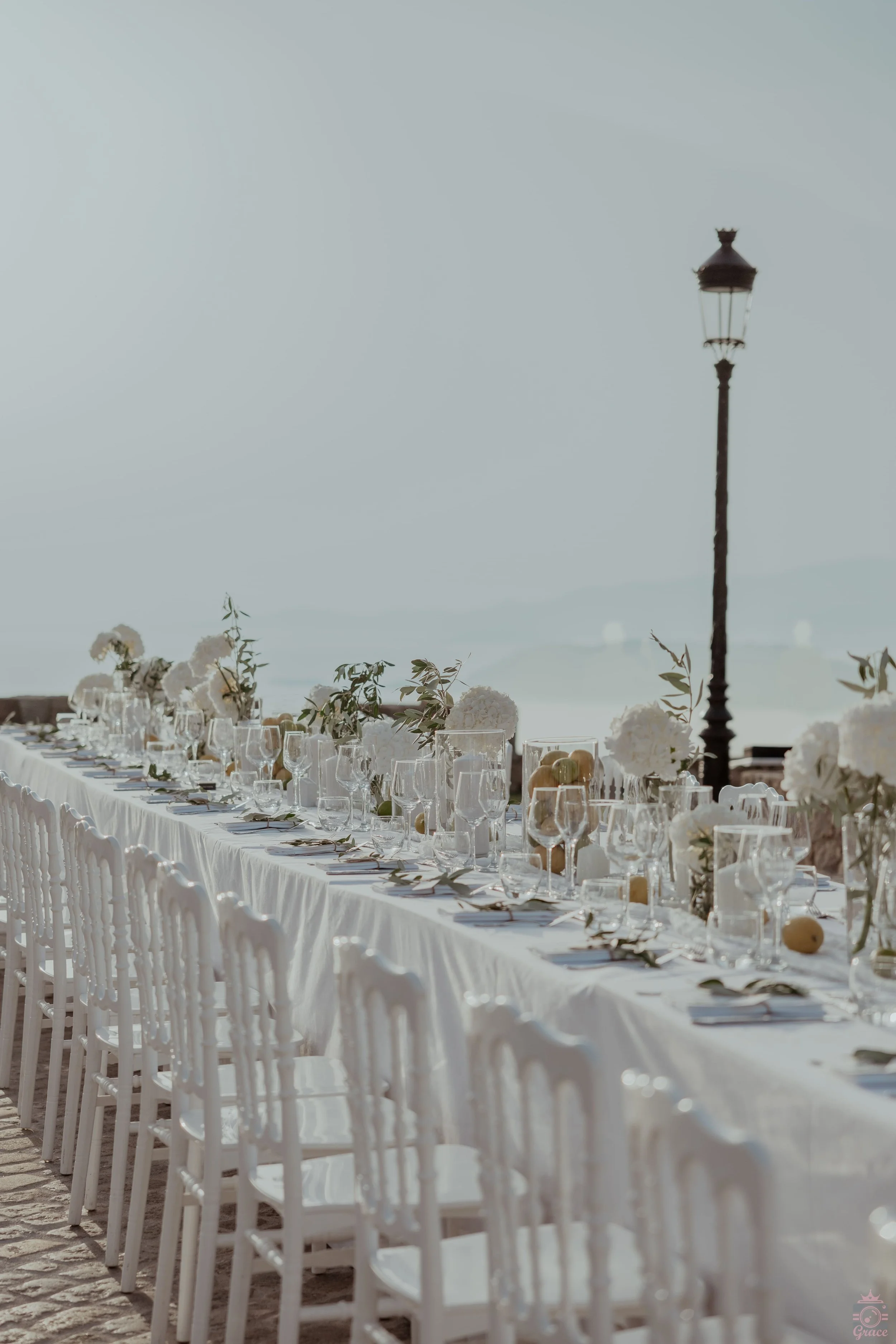Une longue table dressée pour un repas en plein air, décorée avec des fleurs blanches, des verres et des assiettes, sous une lampe antique, avec une mer en arrière-plan.
