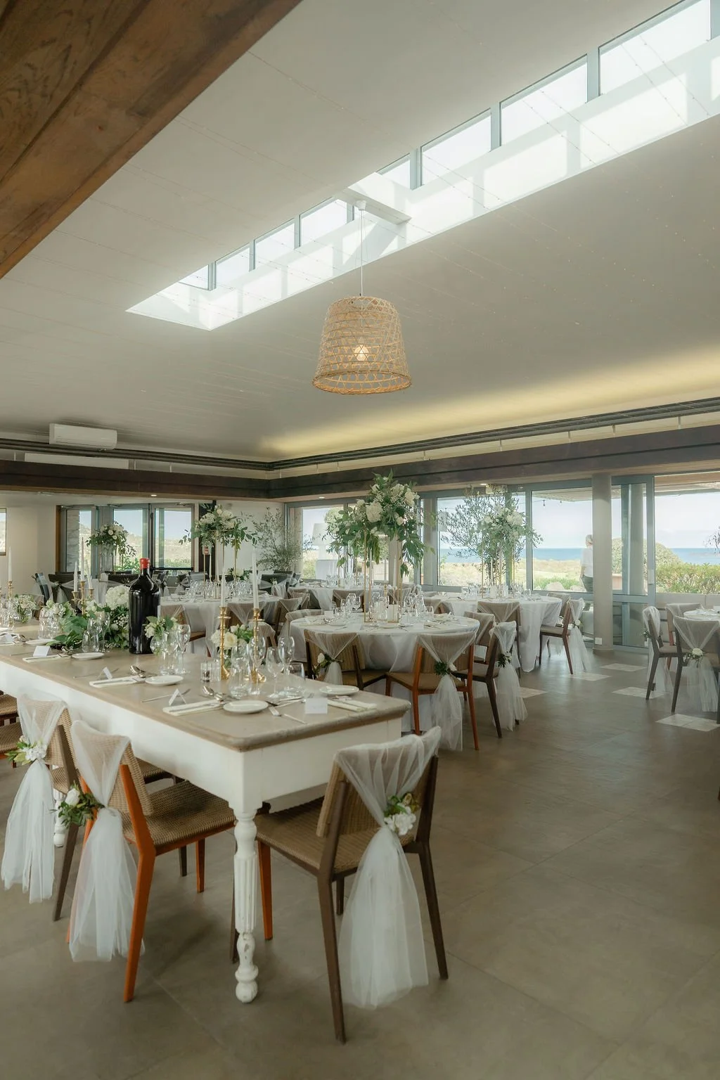 Salle de réception élégante avec tables rondes et décor floral, vue sur la mer à travers de grandes fenêtres, éclairage naturel, décoration raffinée pour un événement spécial.