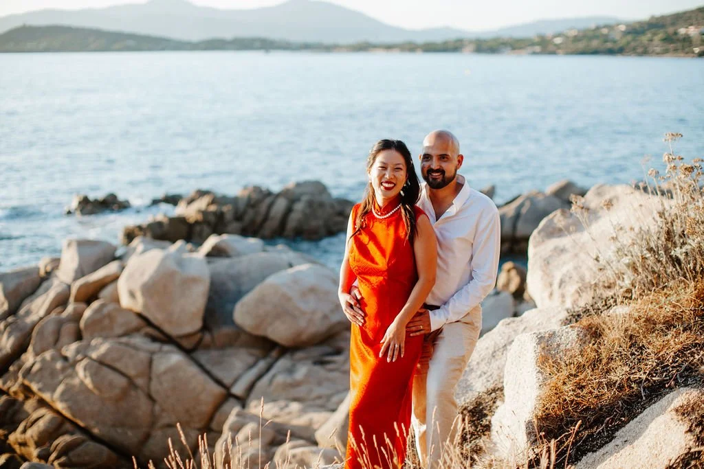 Un couple souriant se tient près de la mer, entouré de rochers et de végétation sèche, avec un paysage de collines en arrière-plan.