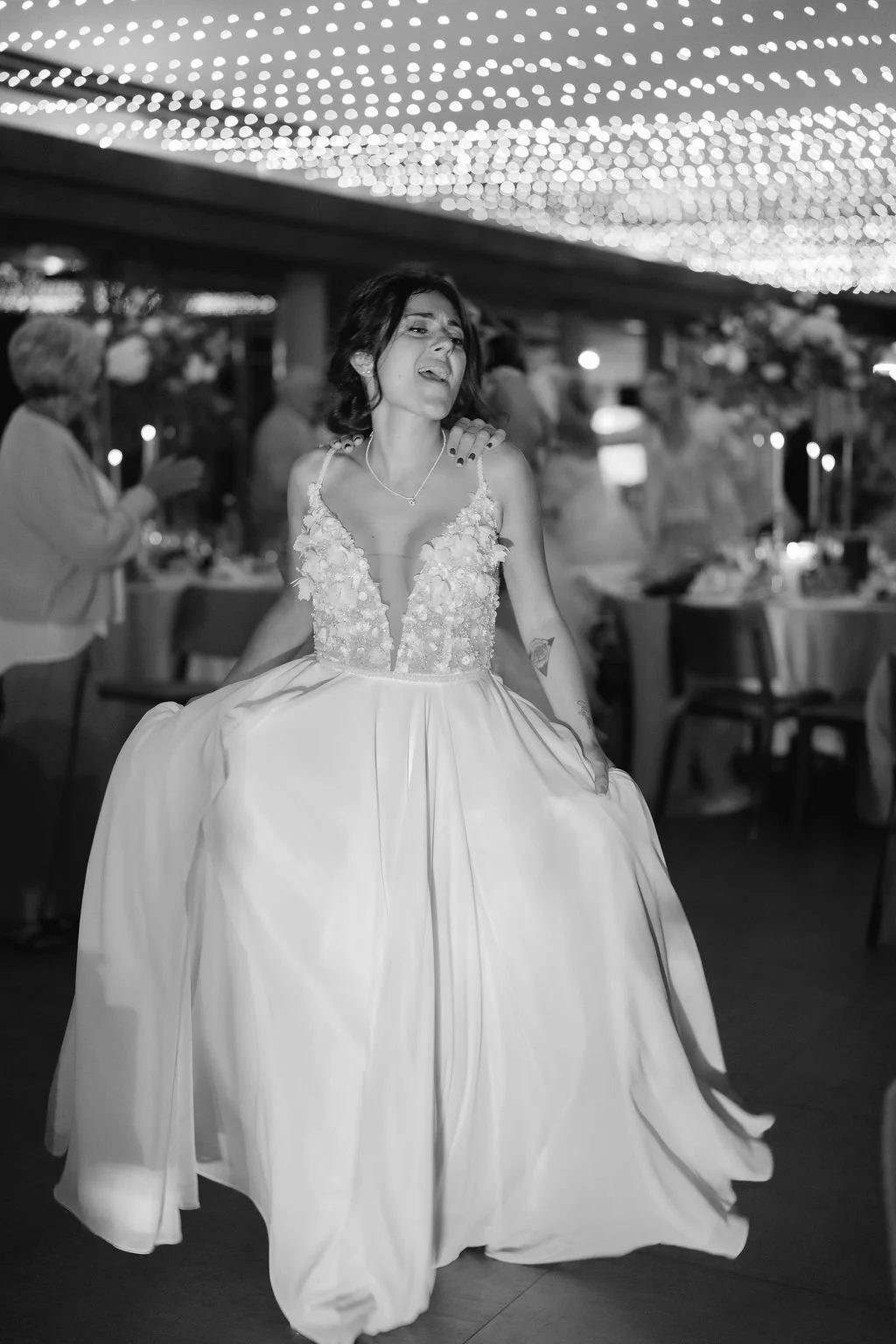 Une femme en robe de mariée danse lors d'une célébration, avec d'autres personnes en arrière-plan dans une salle décorée avec des lumières suspendues.