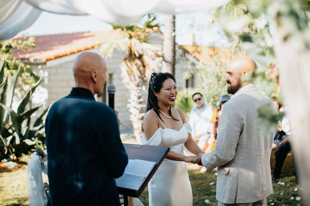 Un couple se marie lors d'une cérémonie en plein air, avec un officiant en train de lire ses vœux et le couple se tenant la main, entourés de plantes et d'amis qui assistent à la cérémonie.