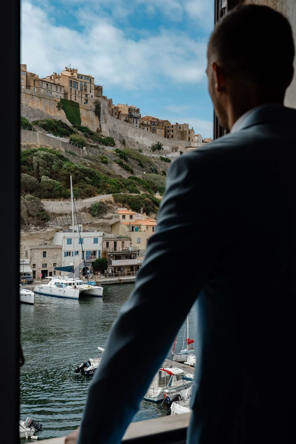 Un homme regarde un port avec des bateaux, un village en hauteur avec des bâtiments en pierre et un ciel bleu avec quelques nuages.