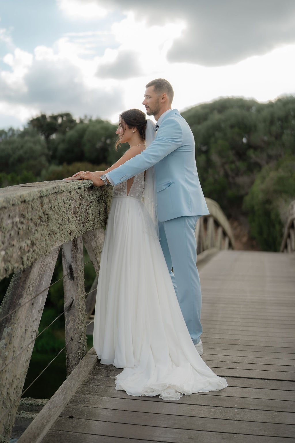 Un couple en tenue de mariage, un homme en costume bleu clair et une femme en robe blanche, se tenant sur un pont en bois en pleine nature, lors d'une journée nuageuse.