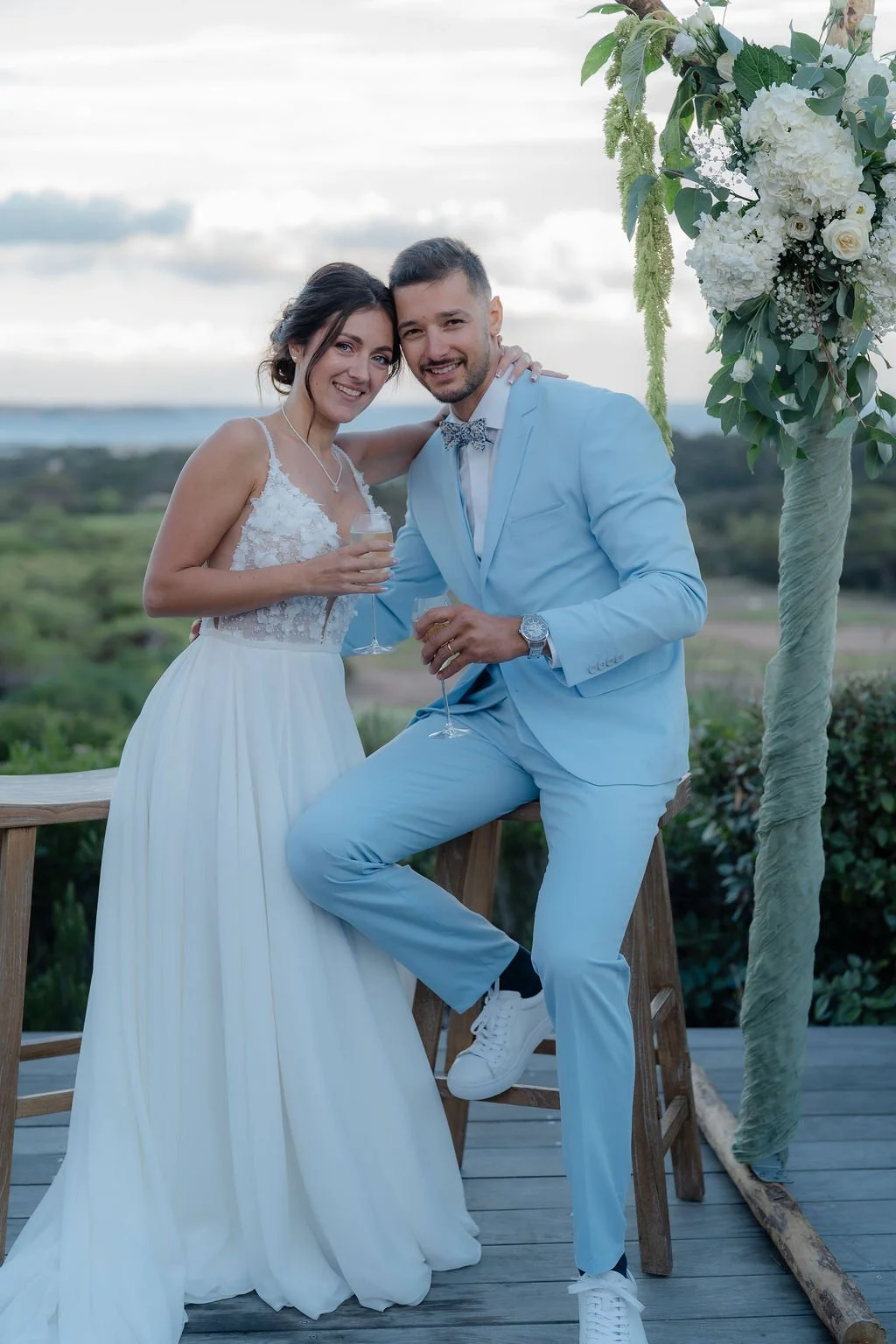 Un couple en vêtements de mariage posant pour une photo lors d'une cérémonie en extérieur, avec un décor naturel en arrière-plan et une arche de fleurs blanche.