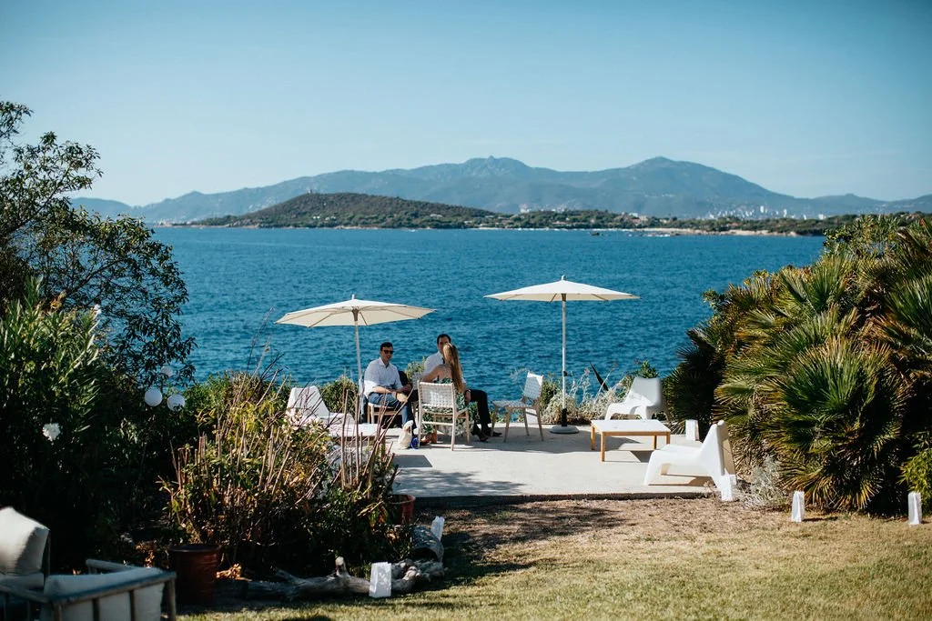 Trois personnes assises sous des parasols blancs près d'une mer, avec des montagnes en arrière-plan, dans un espace extérieur avec des chaises et des plantes green.