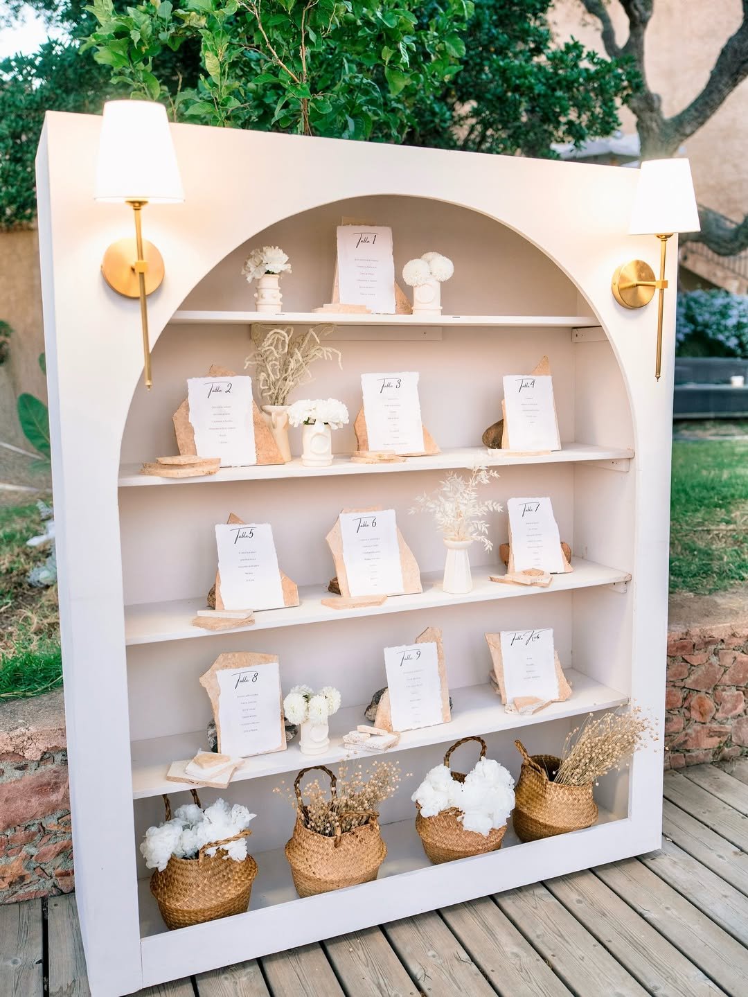 Une étagère blanche décorée de plans de table en forme de pierre, de vases avec fleurs blanches, et de paniers à la base contenant du coton, située en extérieur avec des plantes vertes en arrière-plan.