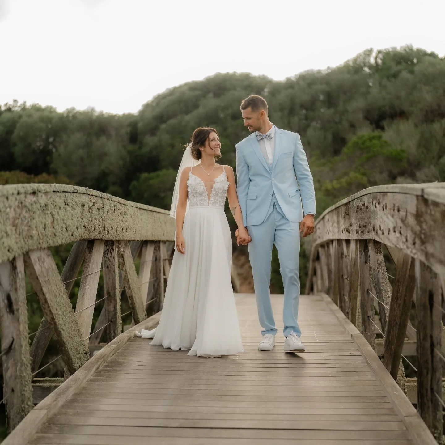 Un couple de mariés se tenant la main sur un pont en bois, avec la mariée en robe blanche et le marié en costume bleu clair, en plein air avec des arbres en arrière-plan.