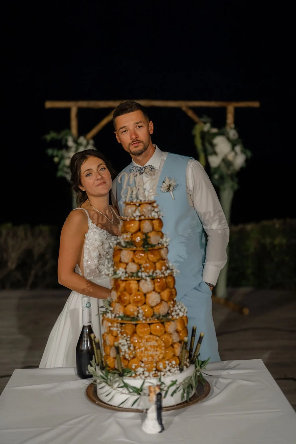 Un couple de mariés posant derrière un grand gâteau de mariage composé de donuts décorés et de petites fleurs.