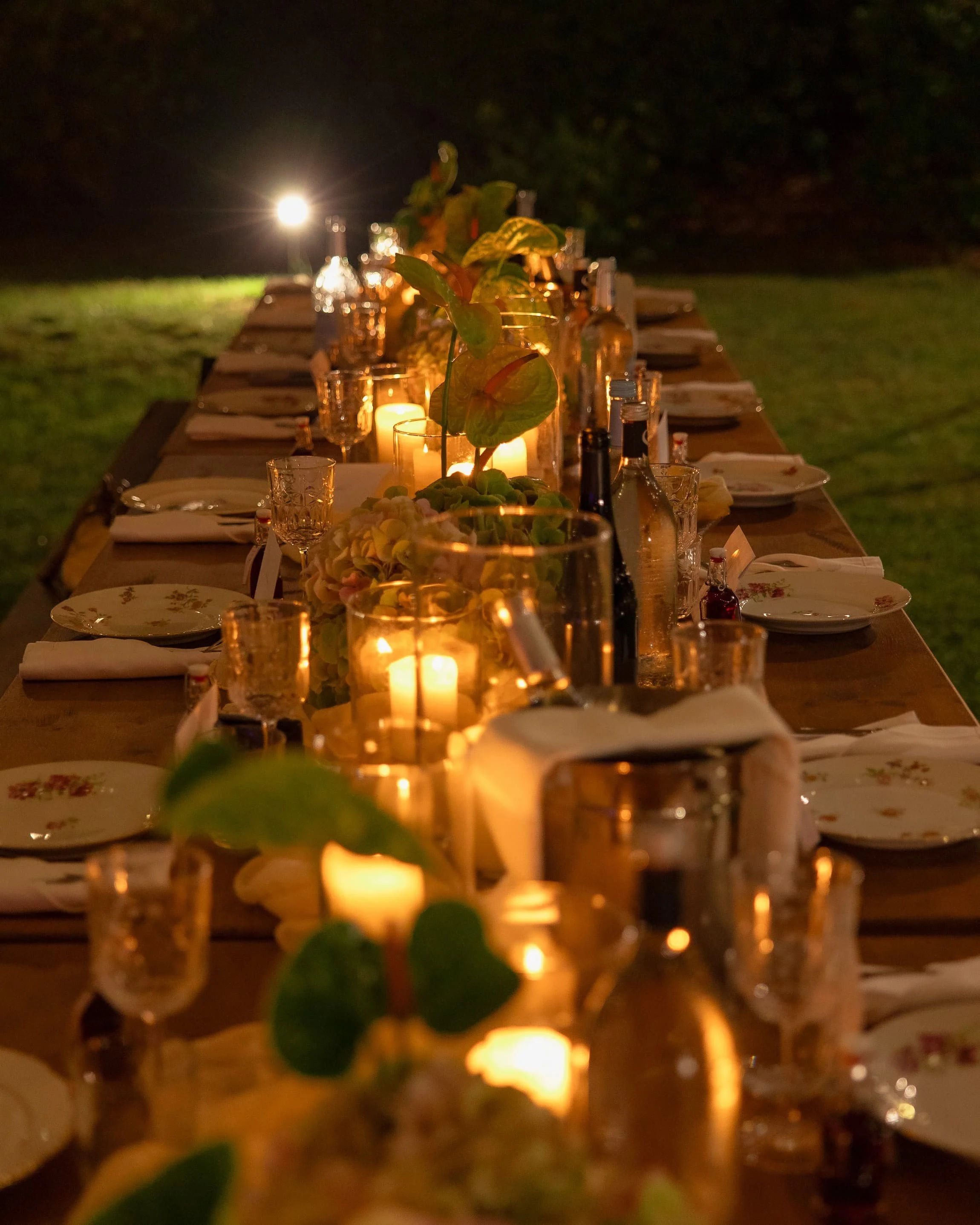 Table de dîner extérieure ornée de bougies, bouteilles et plantes vertes, éclairée tamement, en soirée ou la nuit.