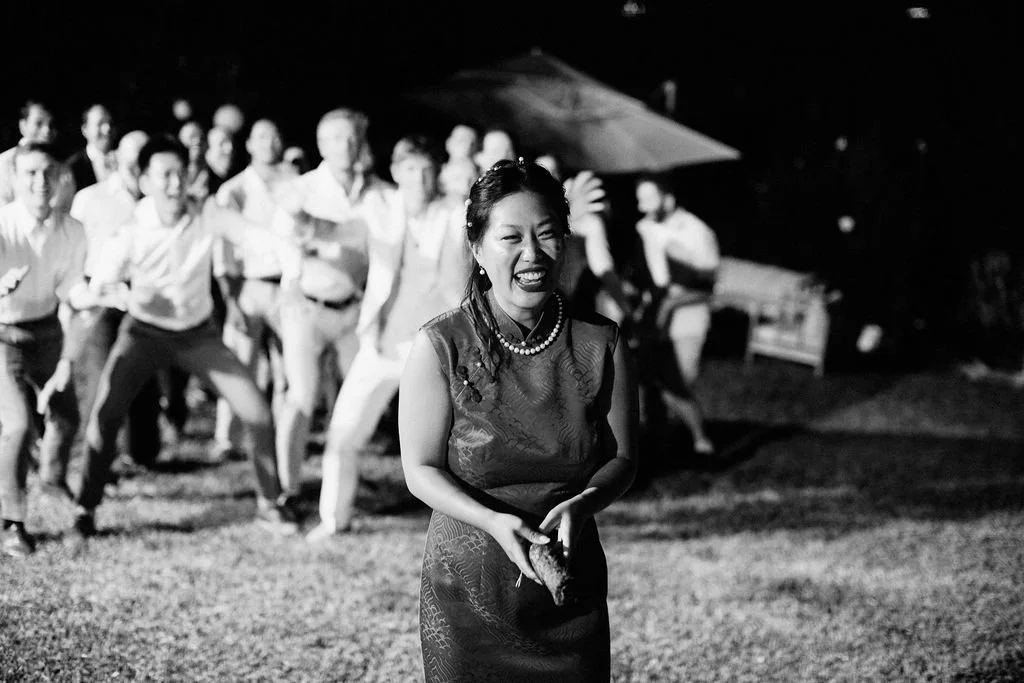Une femme souriante avec un collier de perles au premier plan lors d'une fête en plein air, entourée d'un groupe de personnes dans l'obscurité.