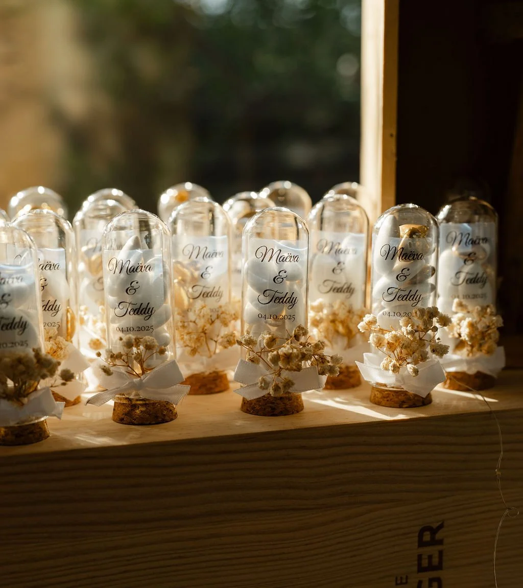 Décor de mariage avec plusieurs bouteilles en verre contenant des pierres et des fleurs, ornées de rubans blanc, sur une table en bois.