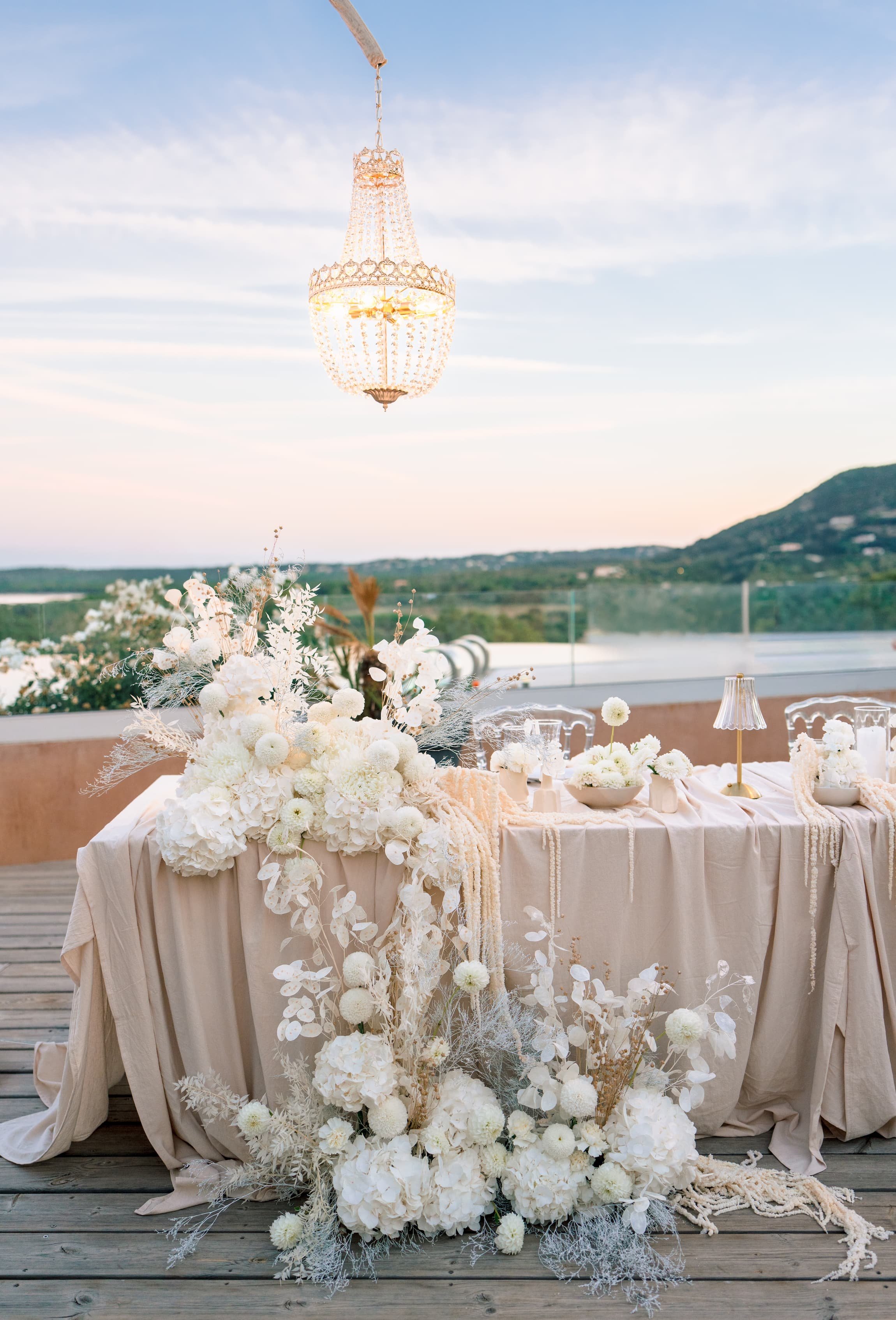 Table de mariage élégante décorée de fleurs blanches, avec un chandelier en cristal suspendu au plafond, sur une terrasse offrant une vue panoramique de la campagne au coucher du soleil.