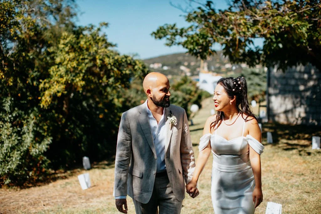 Un couple en tenue de mariage marche main dans la main dans un jardin ensoleillé, entouré d'arbres et de décorations de mariage.