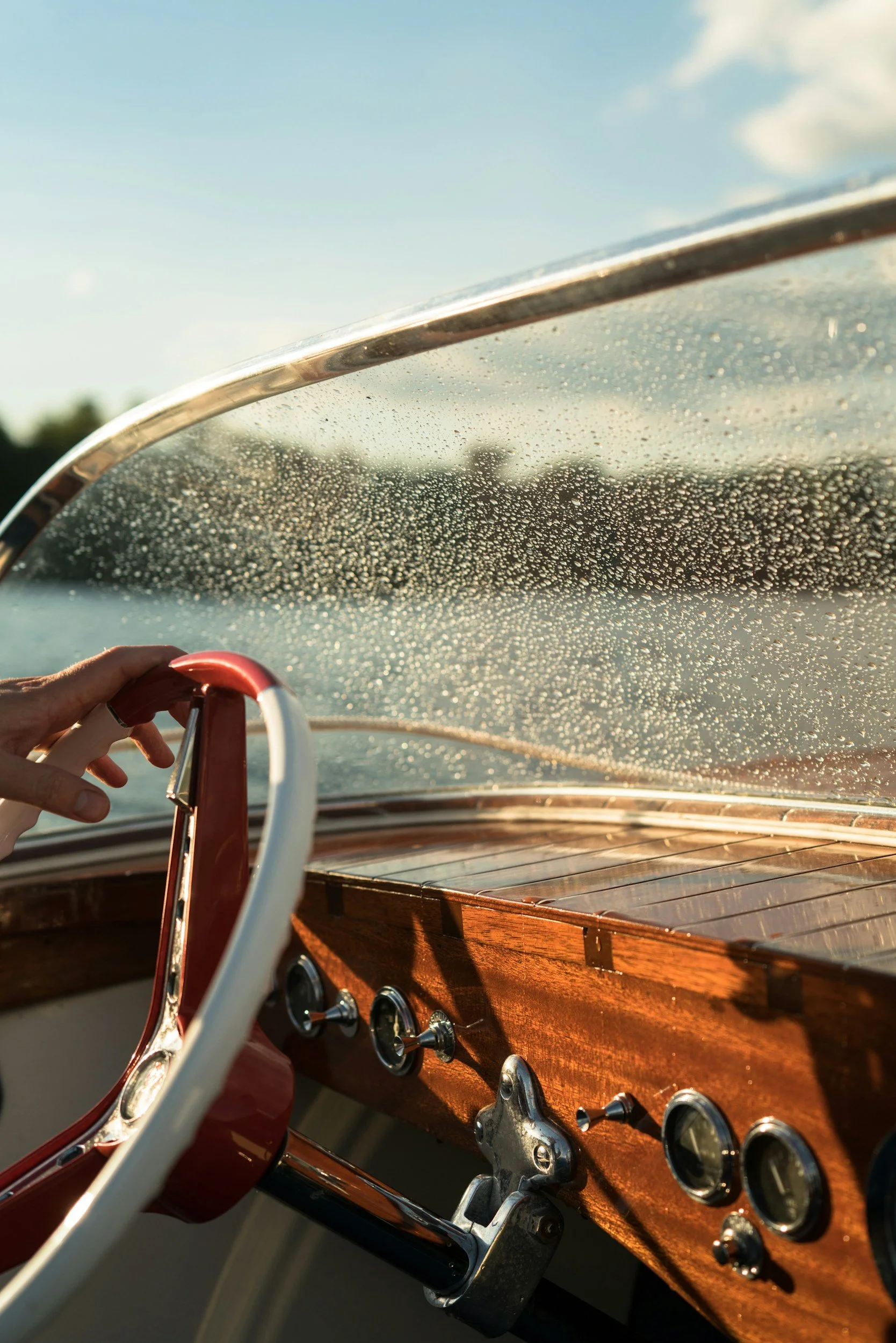 Close-up of a vintage boat's wooden dashboard and steering wheel, with a glass windshield covered in water droplets and a blurred landscape with a blue sky in the background.