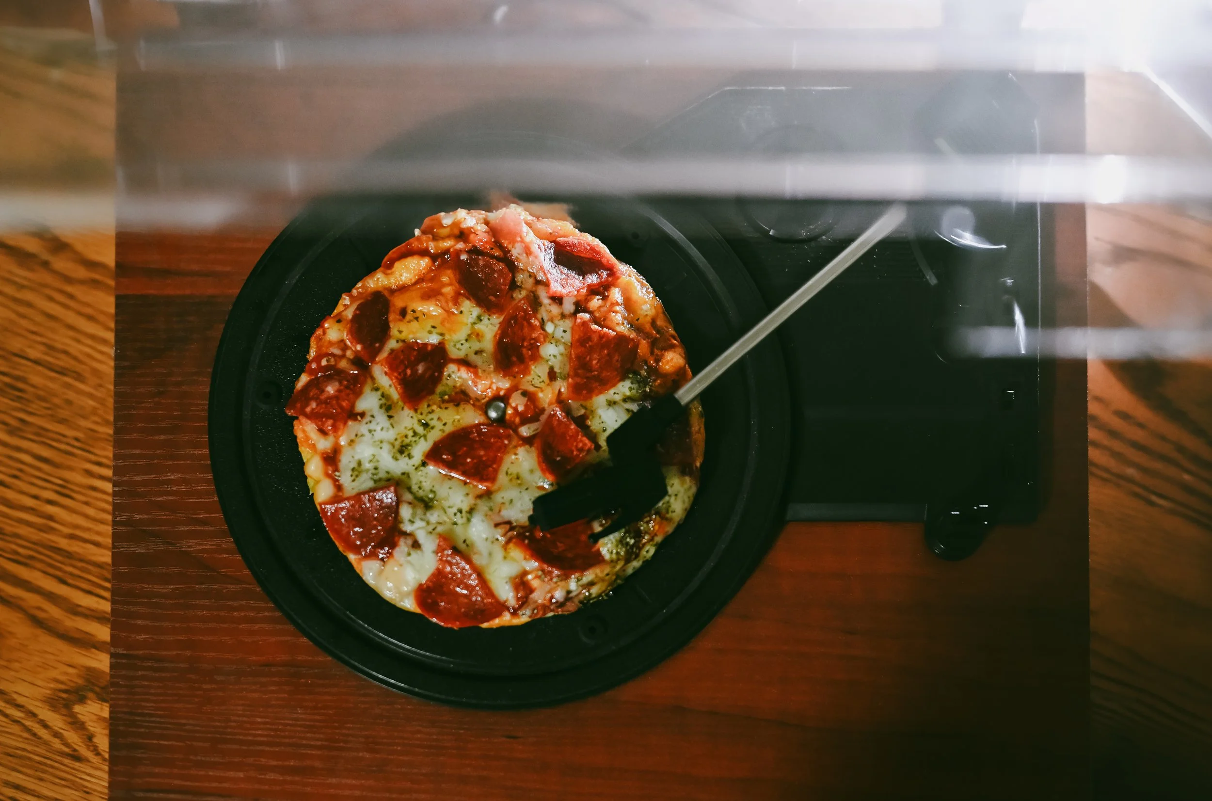 An overhead view of a pepperoni pizza on a black pizza pan, with a pizza cutter resting on top, seen through a transparent cover on a wooden table.