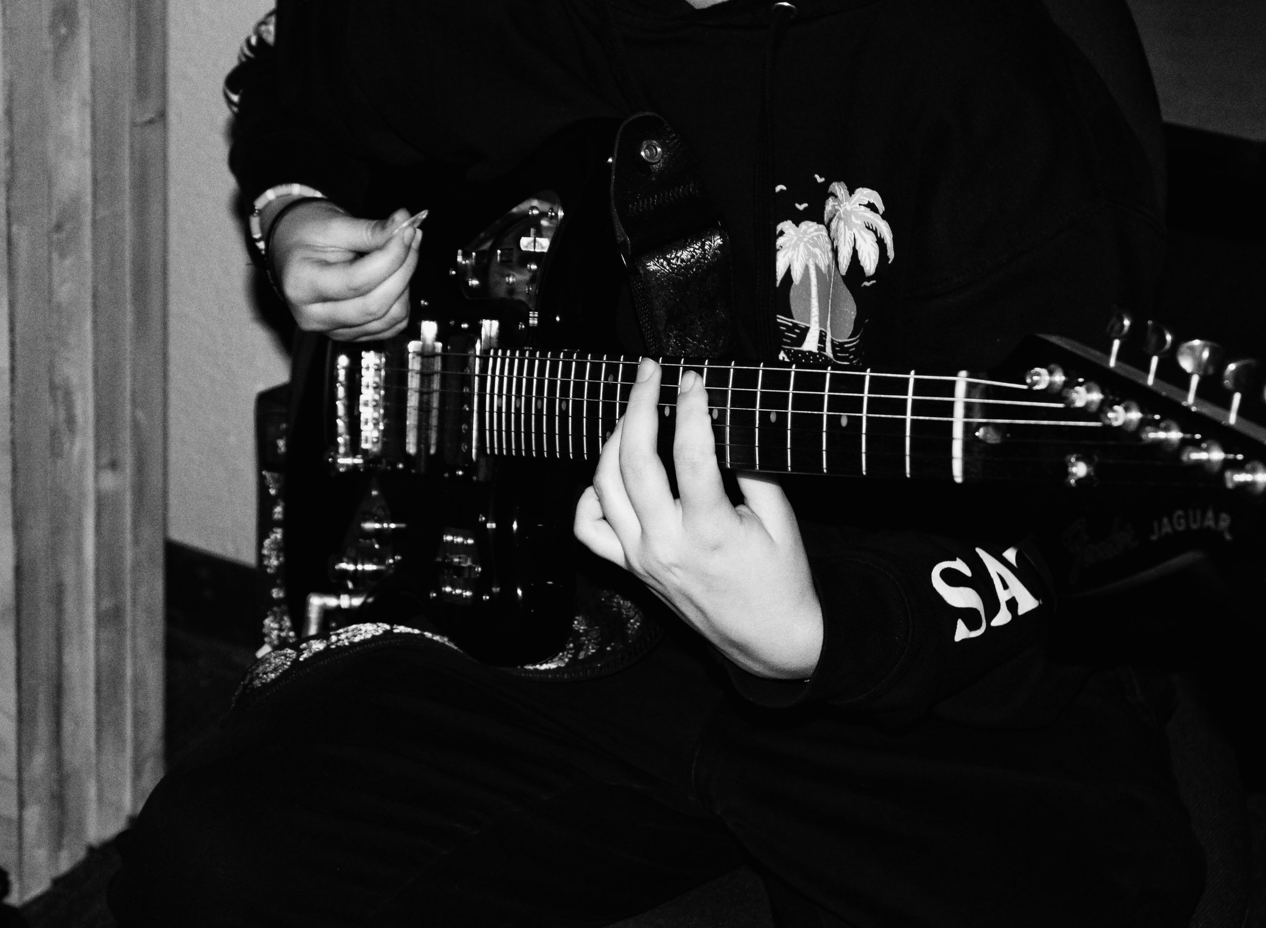 Person playing an electric guitar, wearing a black jacket with a palm tree graphic, sitting on a chair.