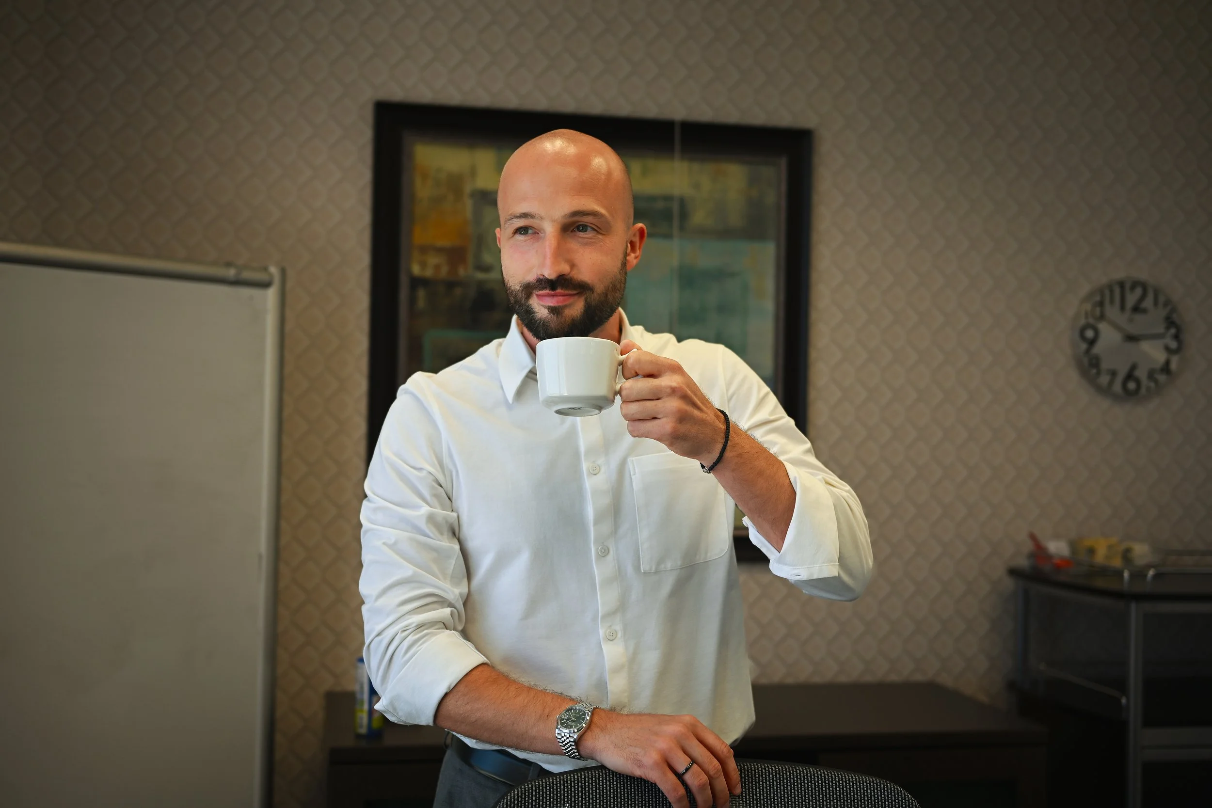 A man in a white shirt holds a coffee mug, standing in an office with patterned wallpaper and a clock on the wall.