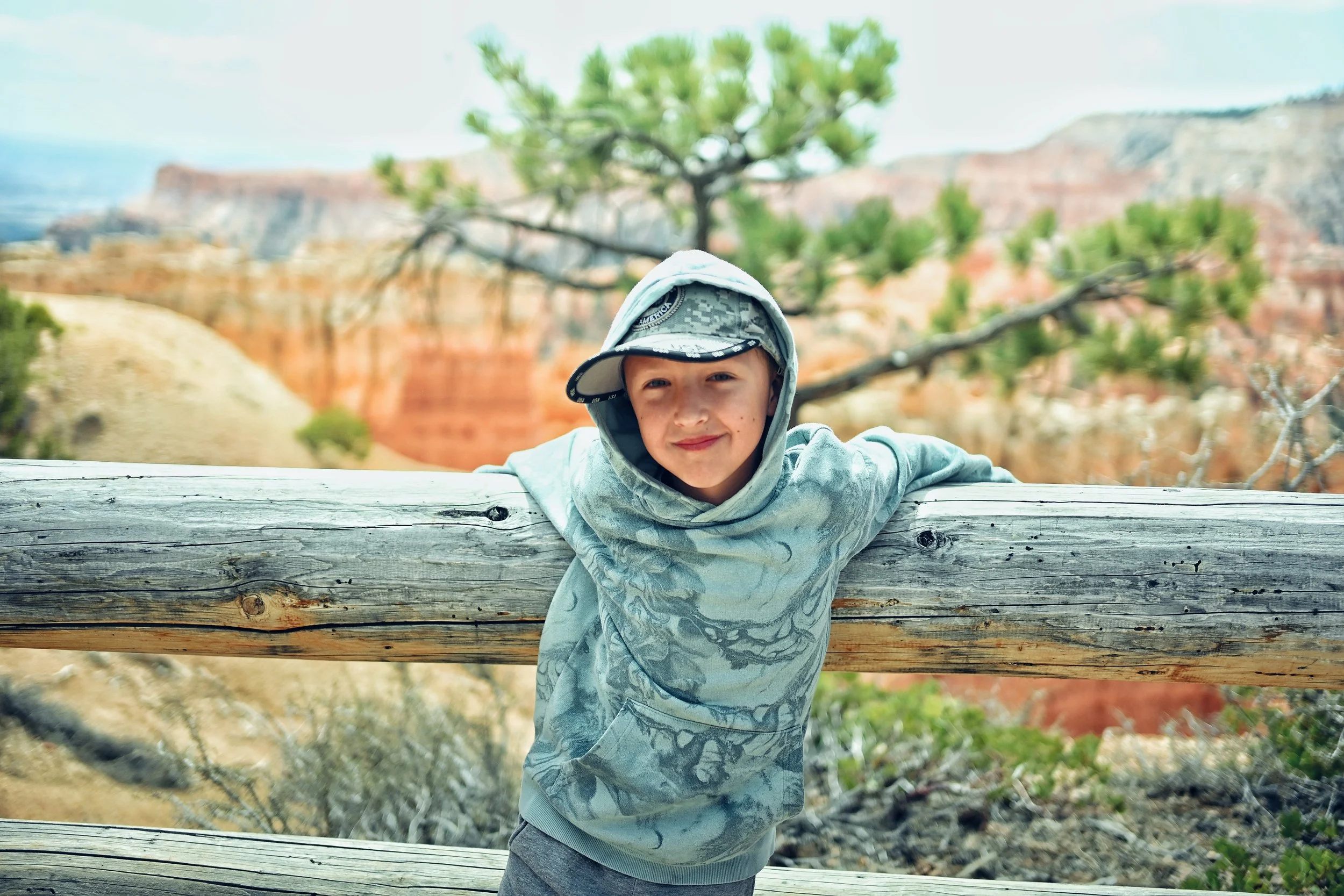 A young boy in a gray hoodie and camouflage cap leaning on a wooden fence, with the Grand Canyon in the background.
