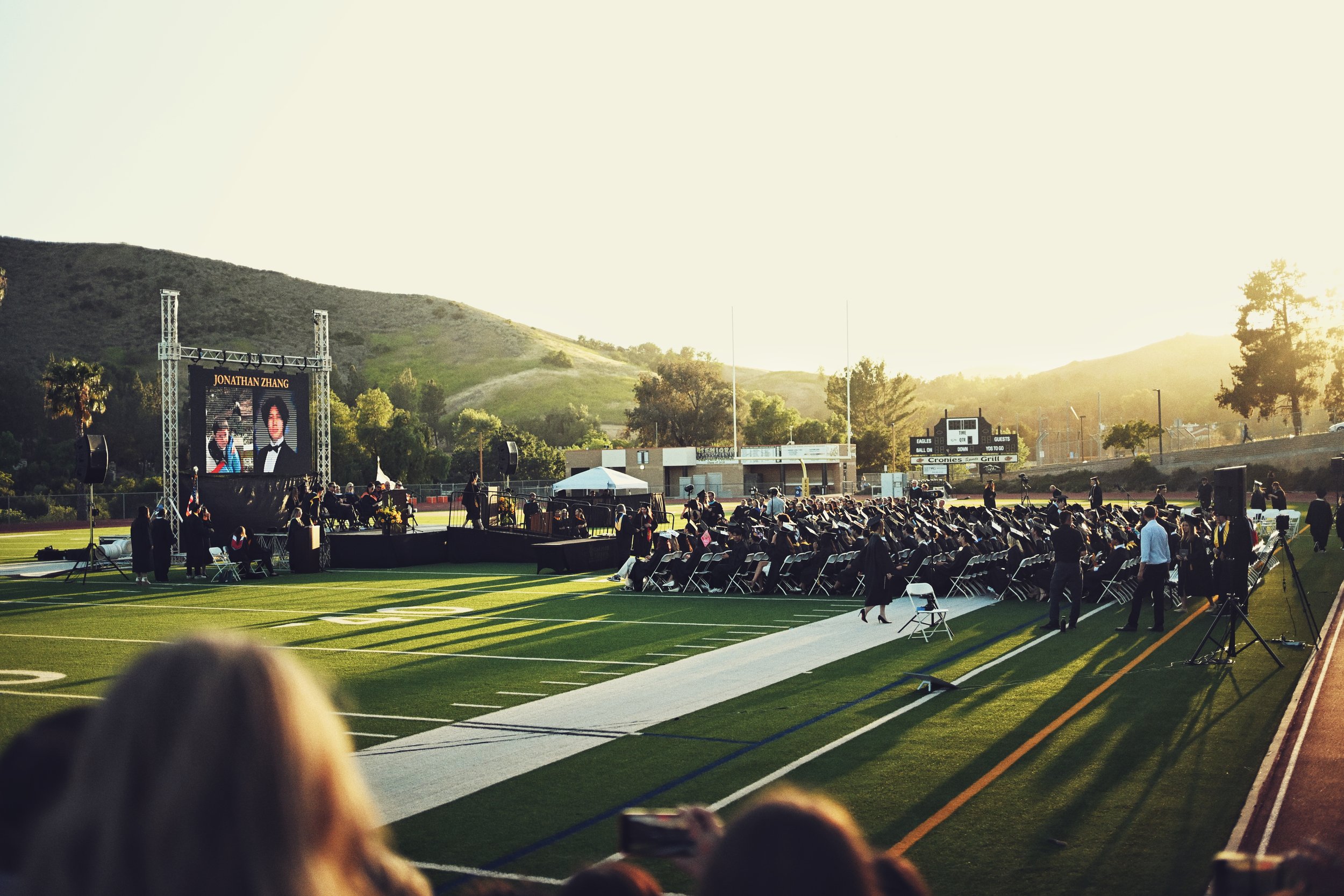 Graduation ceremony at a sports field during sunset with graduates in caps and gowns, seated and facing a stage with a large screen showing a person named Jonathan Zhang, surrounded by family and friends.