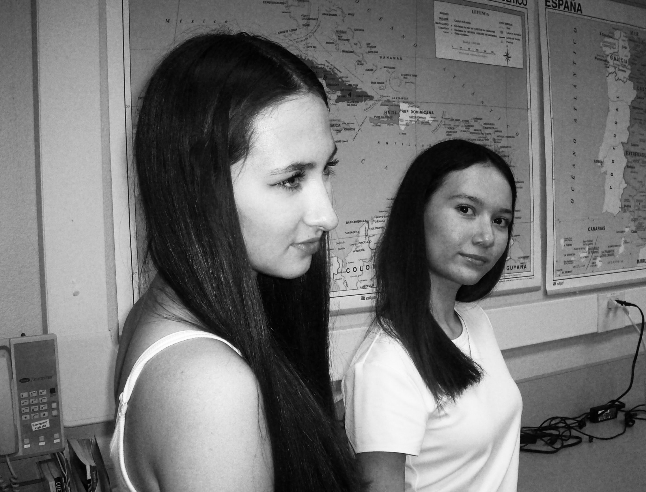 Two young women with long dark hair are standing indoors in front of a wall with maps of Mexico, the Caribbean, and Spain. One is in the foreground, looking thoughtfully to the side; the other is in the background, looking at the camera with a slight smile.