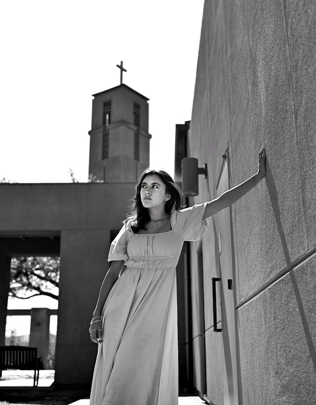 A young woman in a long dress leaning against a textured wall with her arm extended, near a building with a church steeple topped with a cross in the background.