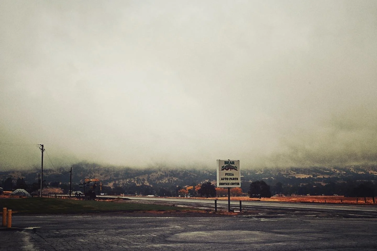 A parking lot with a sign for Bear Mountain Pizza, Auto Parts, and Service Center, with a smoky mountain range in the background under an overcast sky.