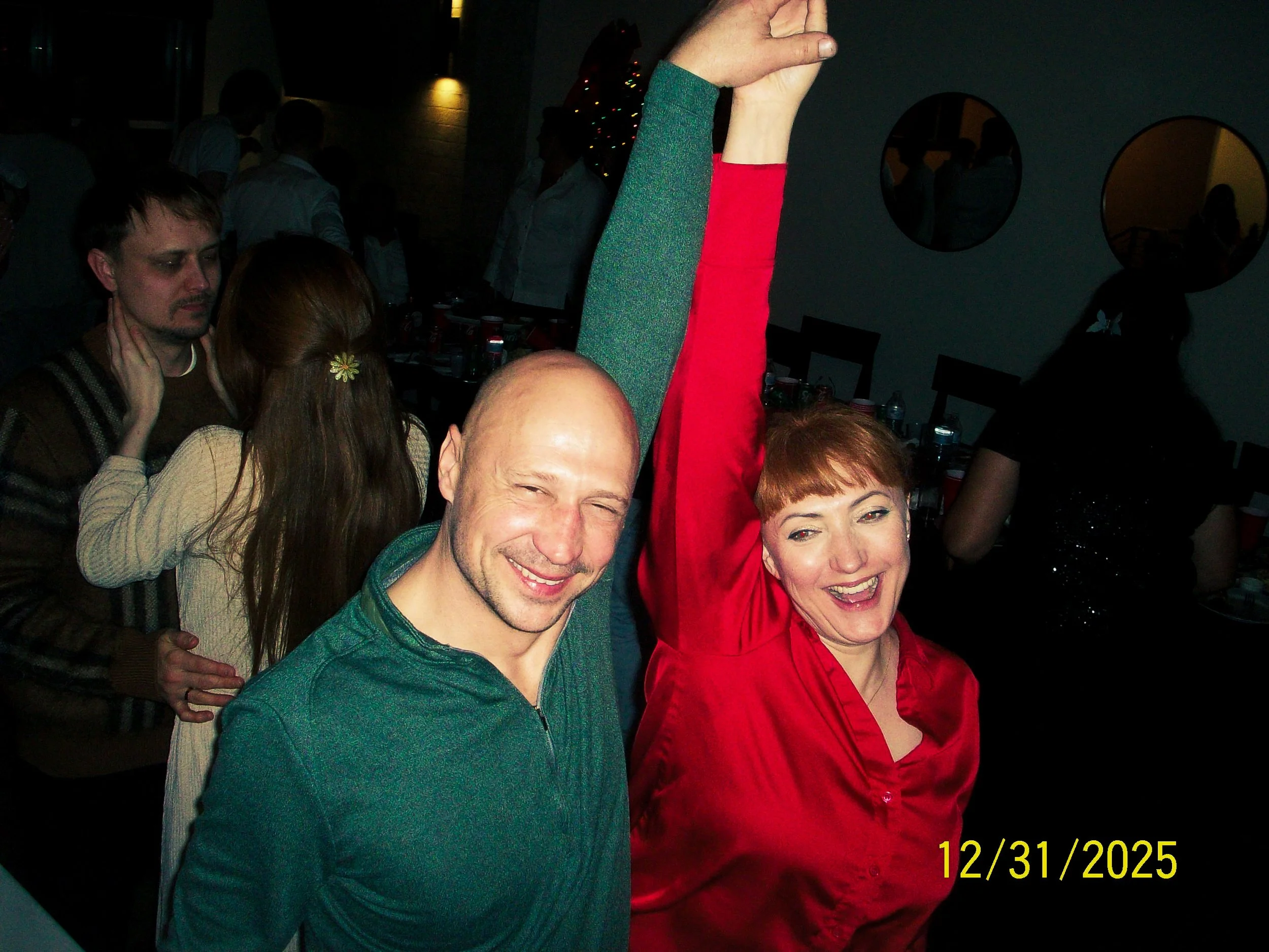 A man and a woman dancing and smiling at a party, with love and joy on their faces. The woman has red hair and is wearing a red top, raising her right arm. The man has a shaved head and is wearing a teal shirt. The background shows other party guests, a decorated Christmas tree, and a dark room with round mirrors on the wall. The date on the photo is December 31, 2025.