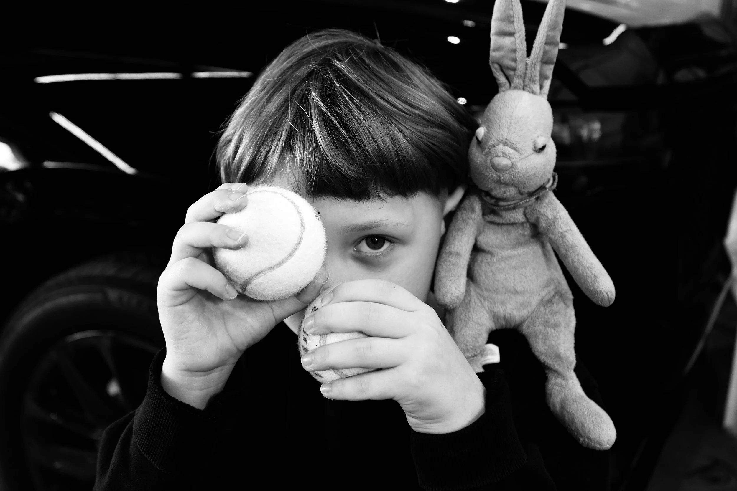 A young boy with short hair holding a tennis ball in front of his face, partly covering one eye, while holding another tennis ball close to his mouth. A soft stuffed rabbit toy is hanging nearby.