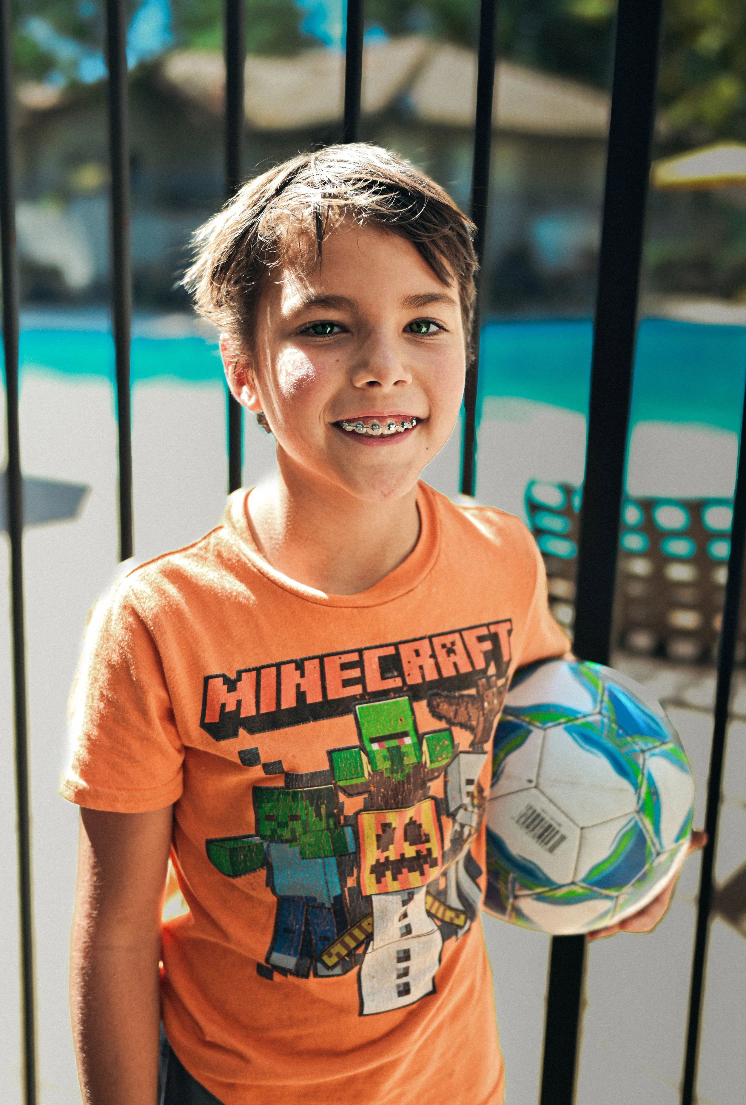 A young boy smiling, holding a colorful soccer ball, standing outdoors near a black metal fence with a swimming pool in the background.