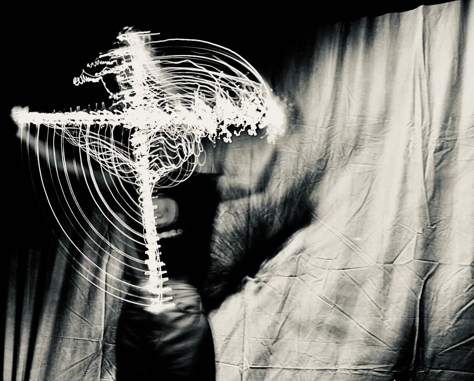 A scientist in gloves holding a clear plastic model of a human brain in front of a large black and white photograph of a person's face with closed eyes. The background is wrinkled fabric.