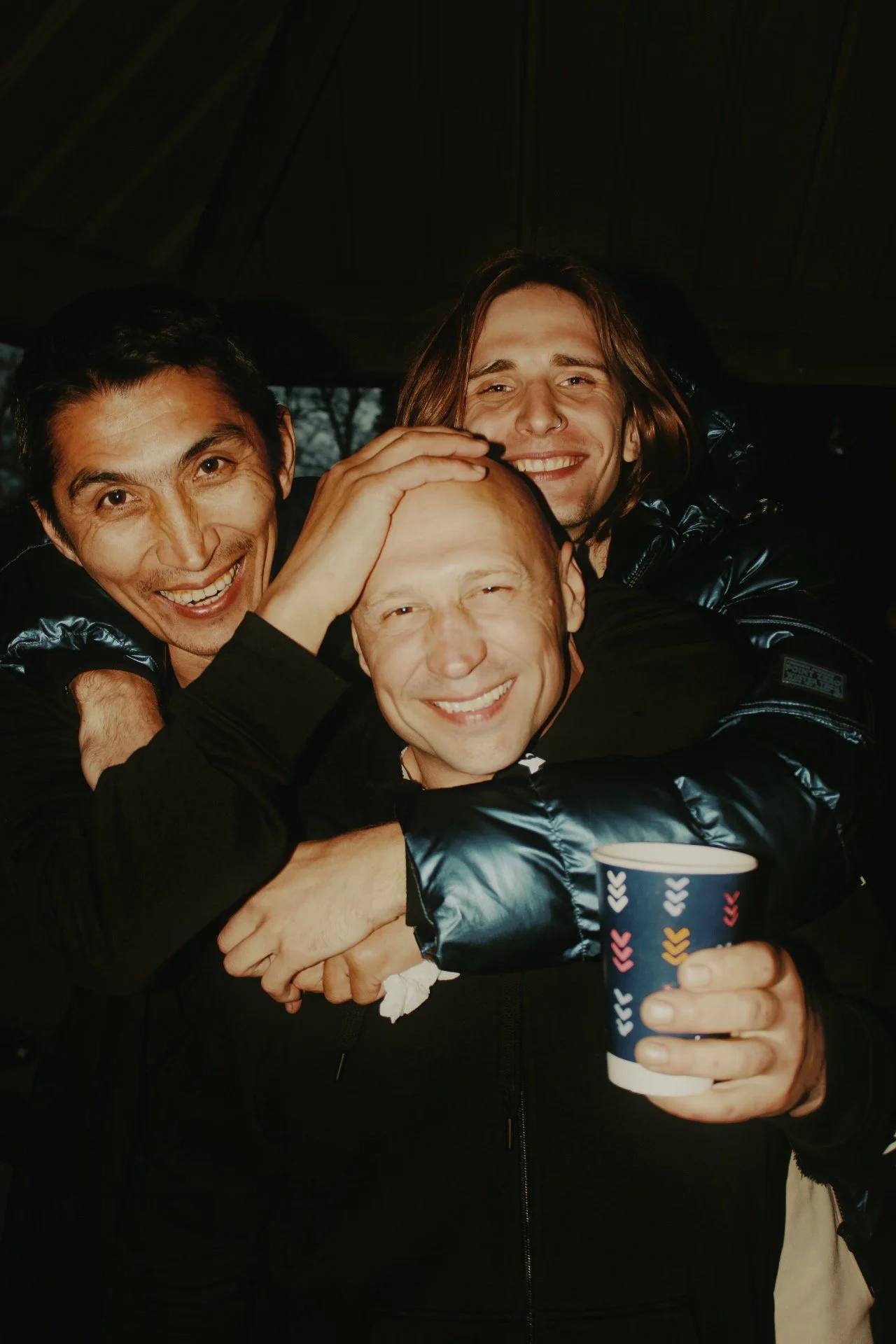 Three men smiling and hugging each other at a gathering, one holding a colorful paper cup.