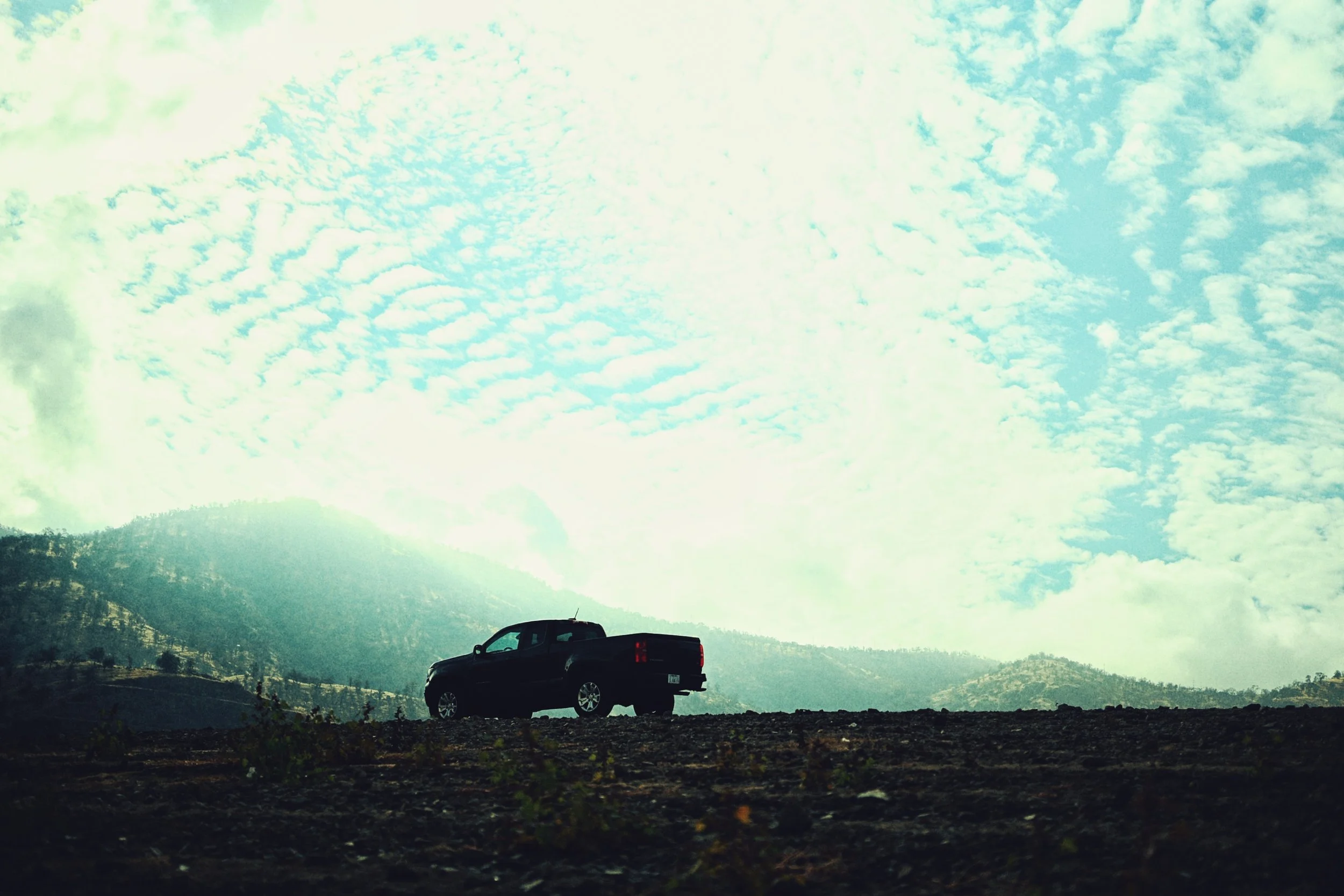 Silhouette of a pickup truck on a dirt road with mountains and a partly cloudy sky in the background