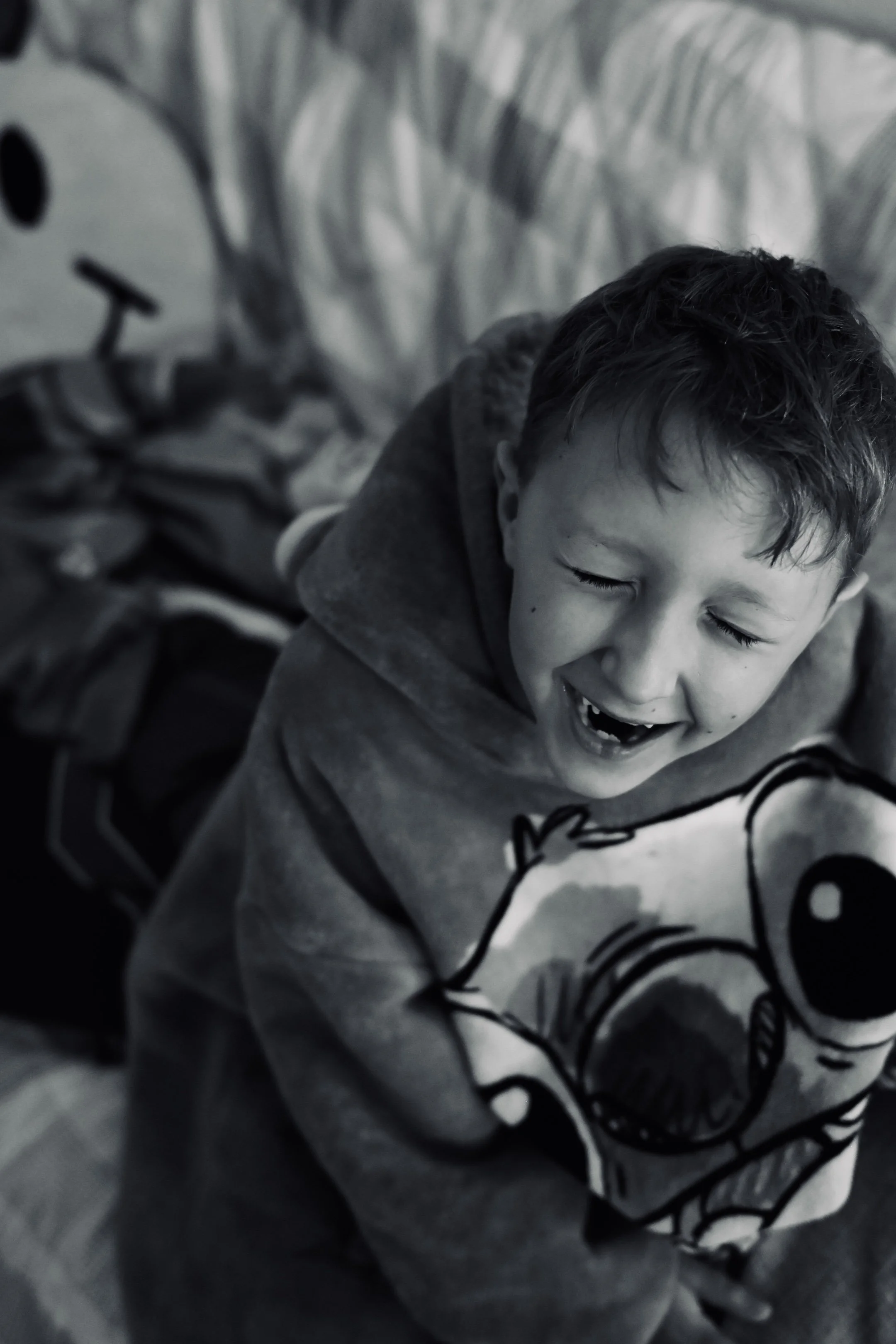 A young boy with short, dark hair smiling with eyes closed, hugging a plush toy with a cartoon dog's face, in a room with striped bedding.