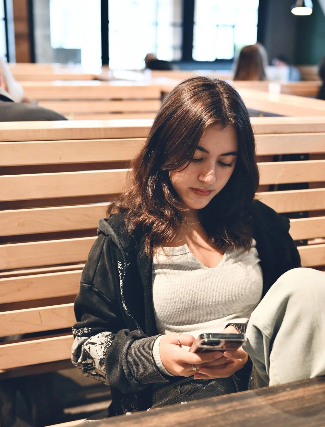 A young woman with shoulder-length dark hair, wearing a light gray top and a black jacket with white patterns on the sleeves, sitting on a wooden bench in a modern, well-lit indoor space, looking at her smartphone.