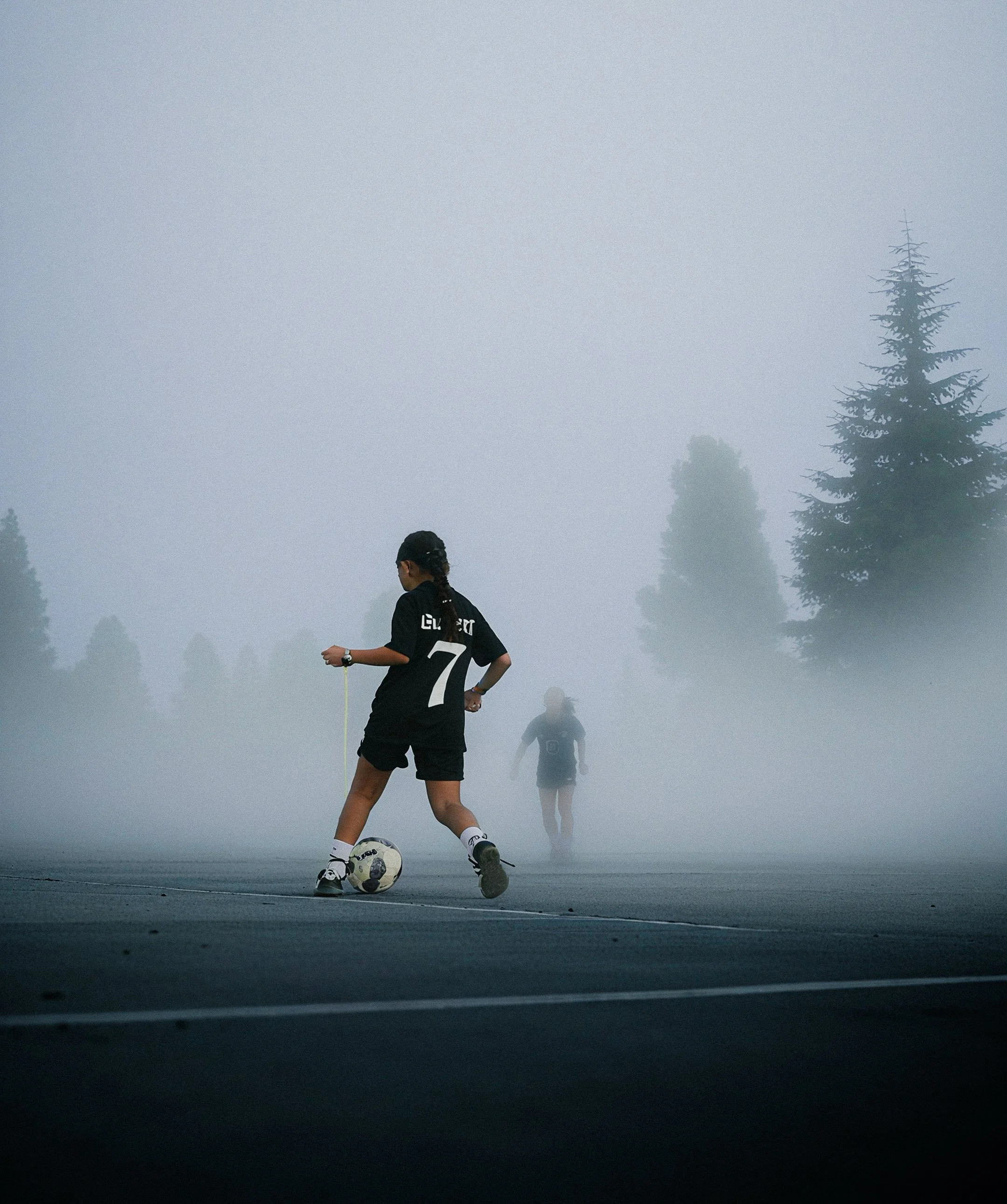 A young girl playing soccer on a foggy outdoor court with two other children in the background.