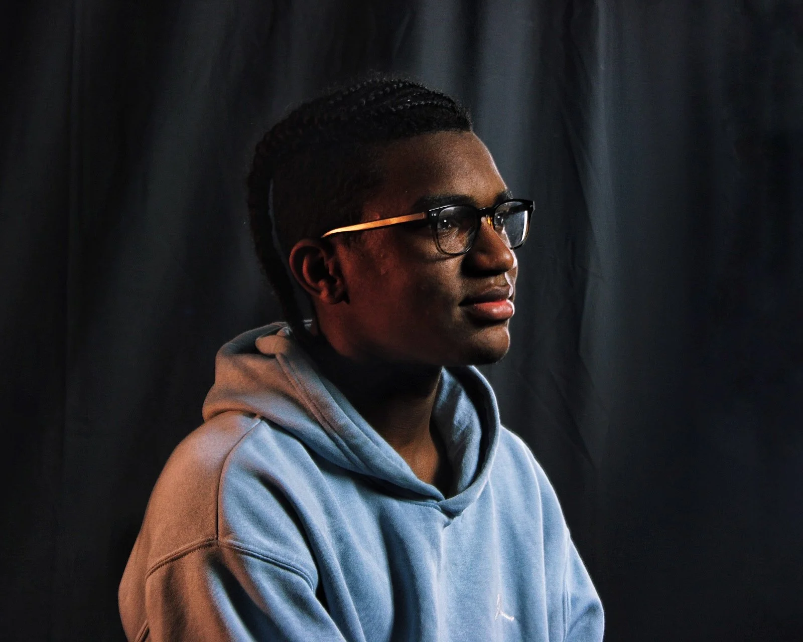 Side profile of a young man with glasses, braided hair, wearing a blue hoodie against a dark background.
