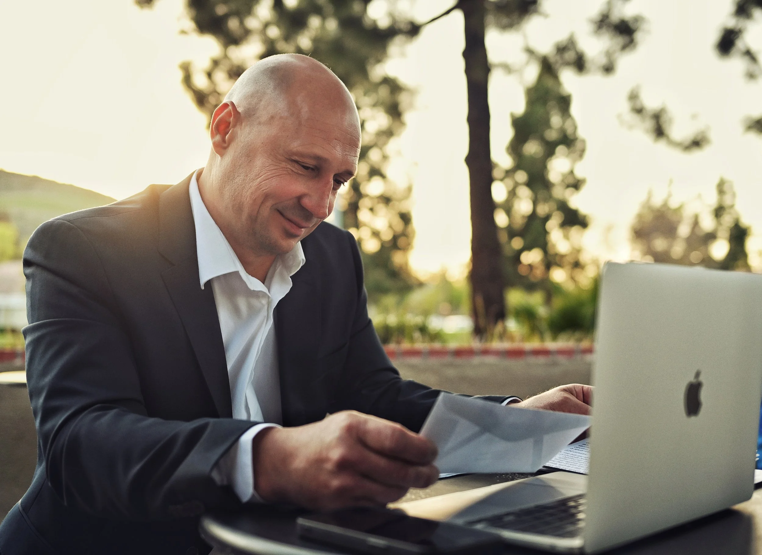 A man in a business suit smiling while working on papers at an outdoor table with a laptop, trees and a sunset in the background.