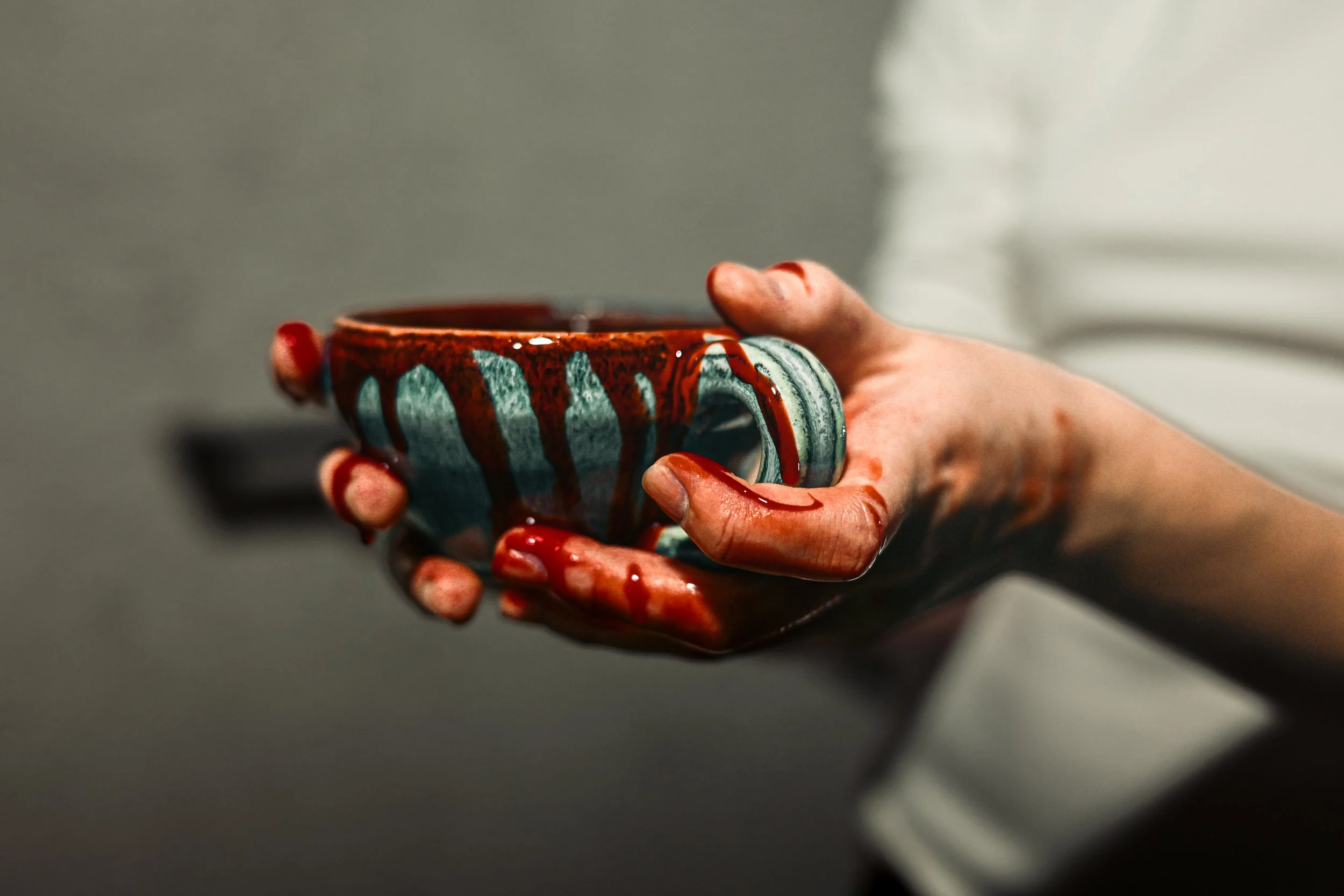 A hand holding a ceramic mug with blue and brown glaze, covered in red paint or liquid. The background is blurred.