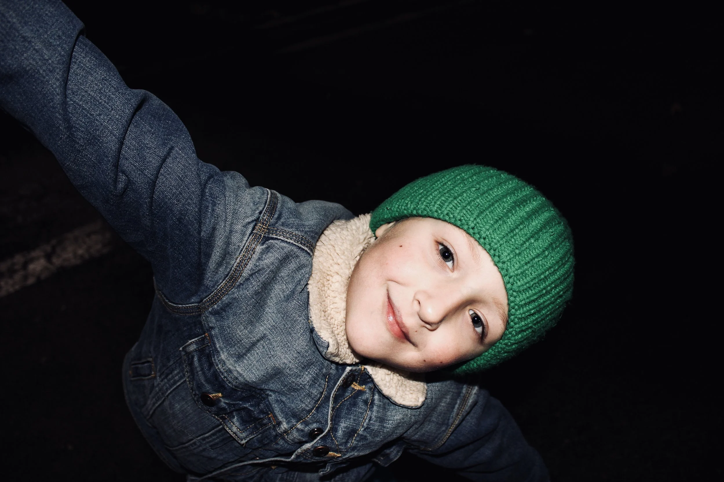 A young child wearing a green knit beanie, denim jacket with a fleece collar, and smiling at the camera against a dark background.